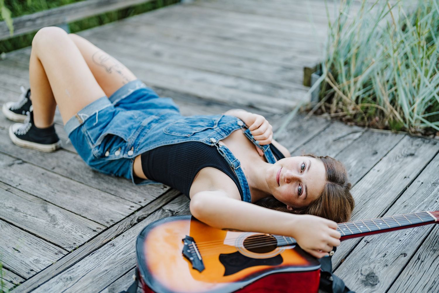 A person in denim shorts lies on a wooden boardwalk with an acoustic guitar in a natural outdoor setting.