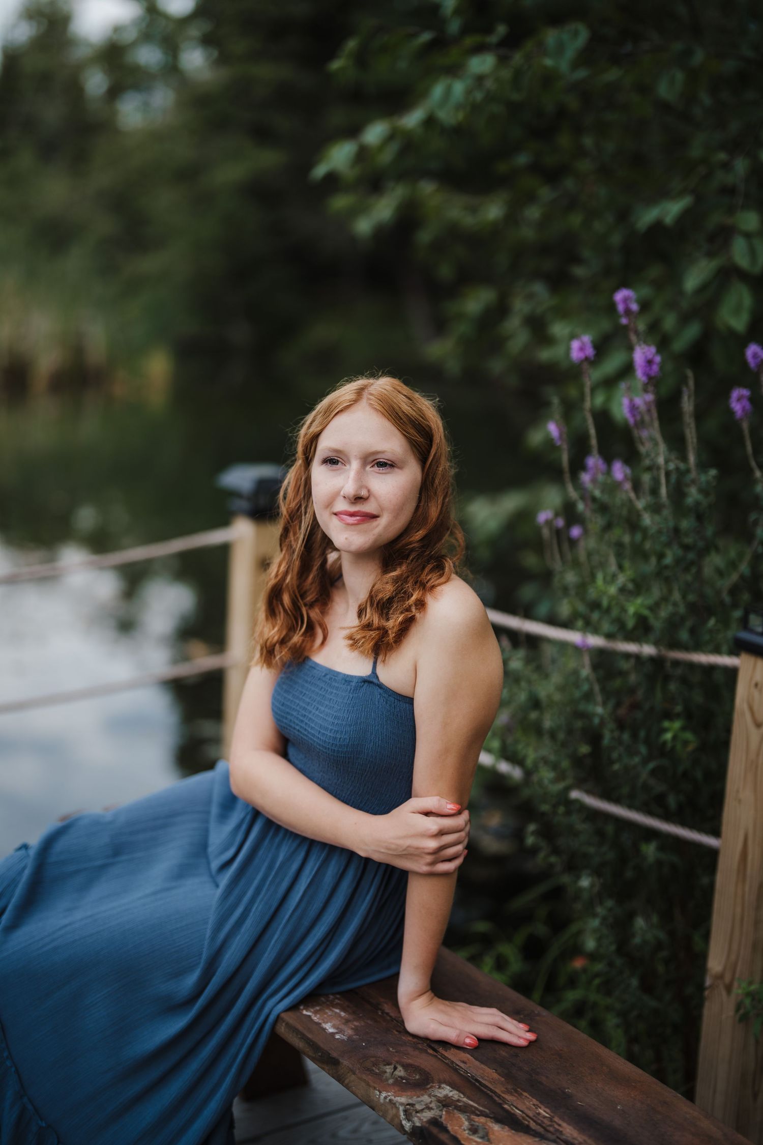 A figure in a navy blue dress poses gracefully near purple wildflowers and a wooden fence by a lake.