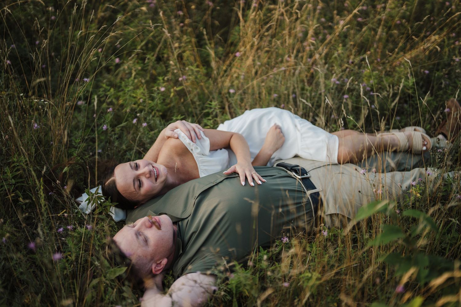 Two people lying together in tall grass during a romantic outdoor photo session at golden hour.