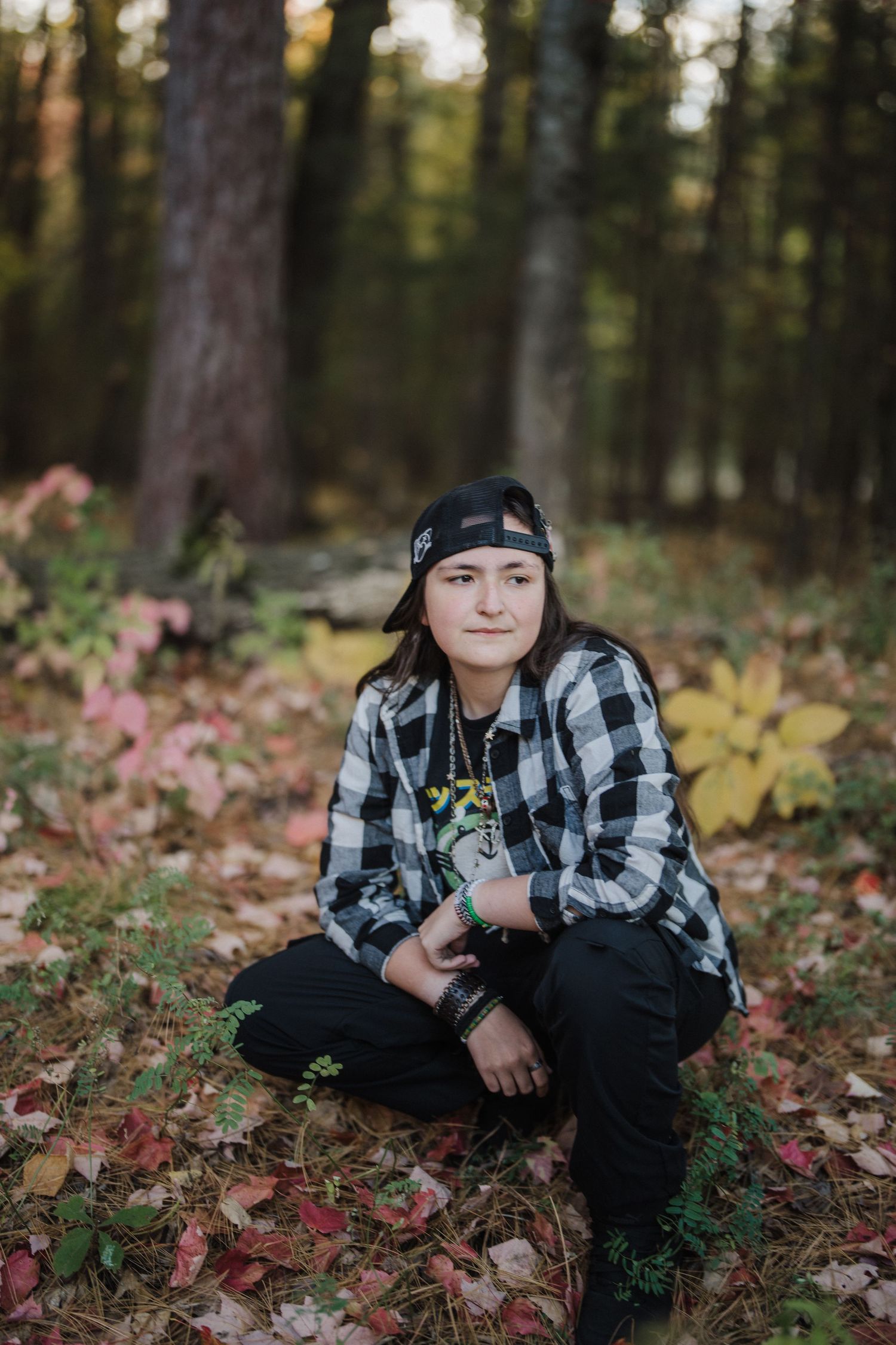 Person wearing plaid flannel shirt and black pants sits among autumn leaves in a wooded forest setting.