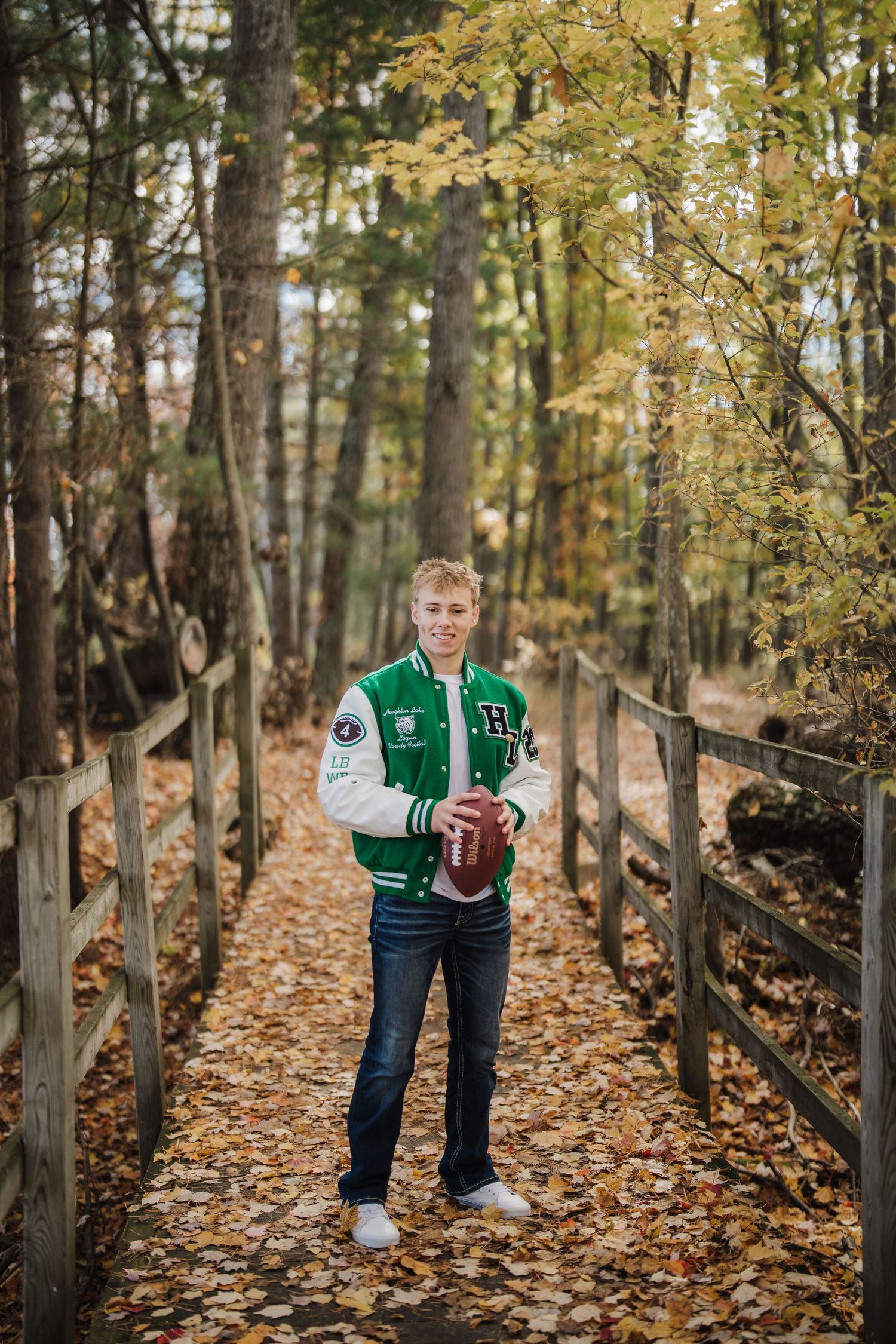 Athletic varsity jacket photo session along a wooden fence path covered in fallen autumn leaves.