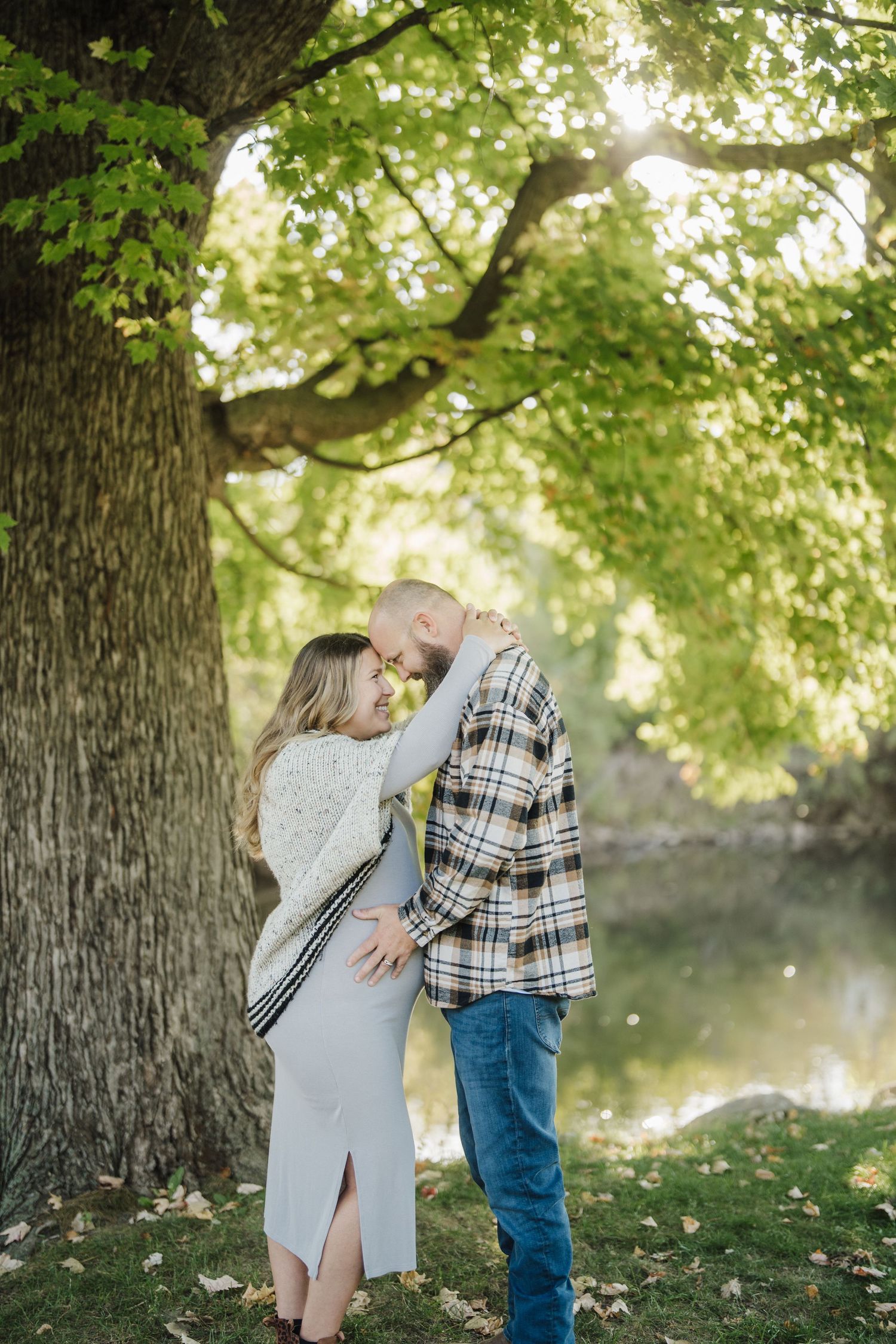Romantic couple shares tender moments under a lush green tree by the water on a sunny afternoon.