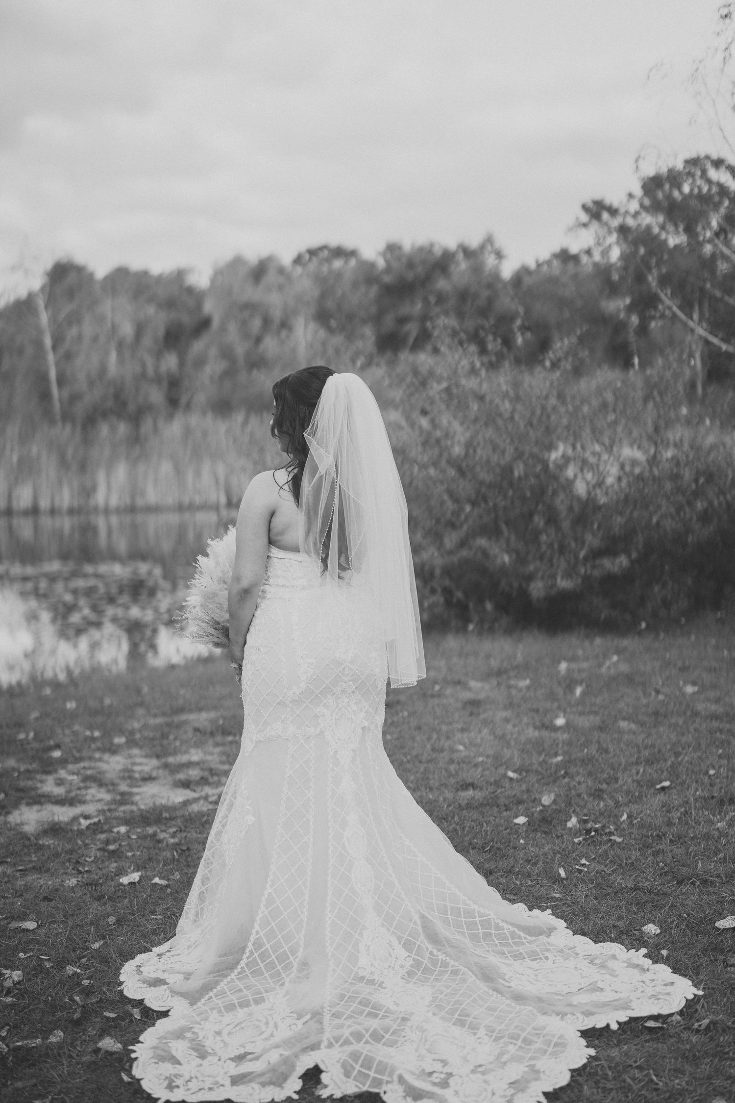 Black and white portrait of bride in lace wedding dress with flowing veil in outdoor setting.