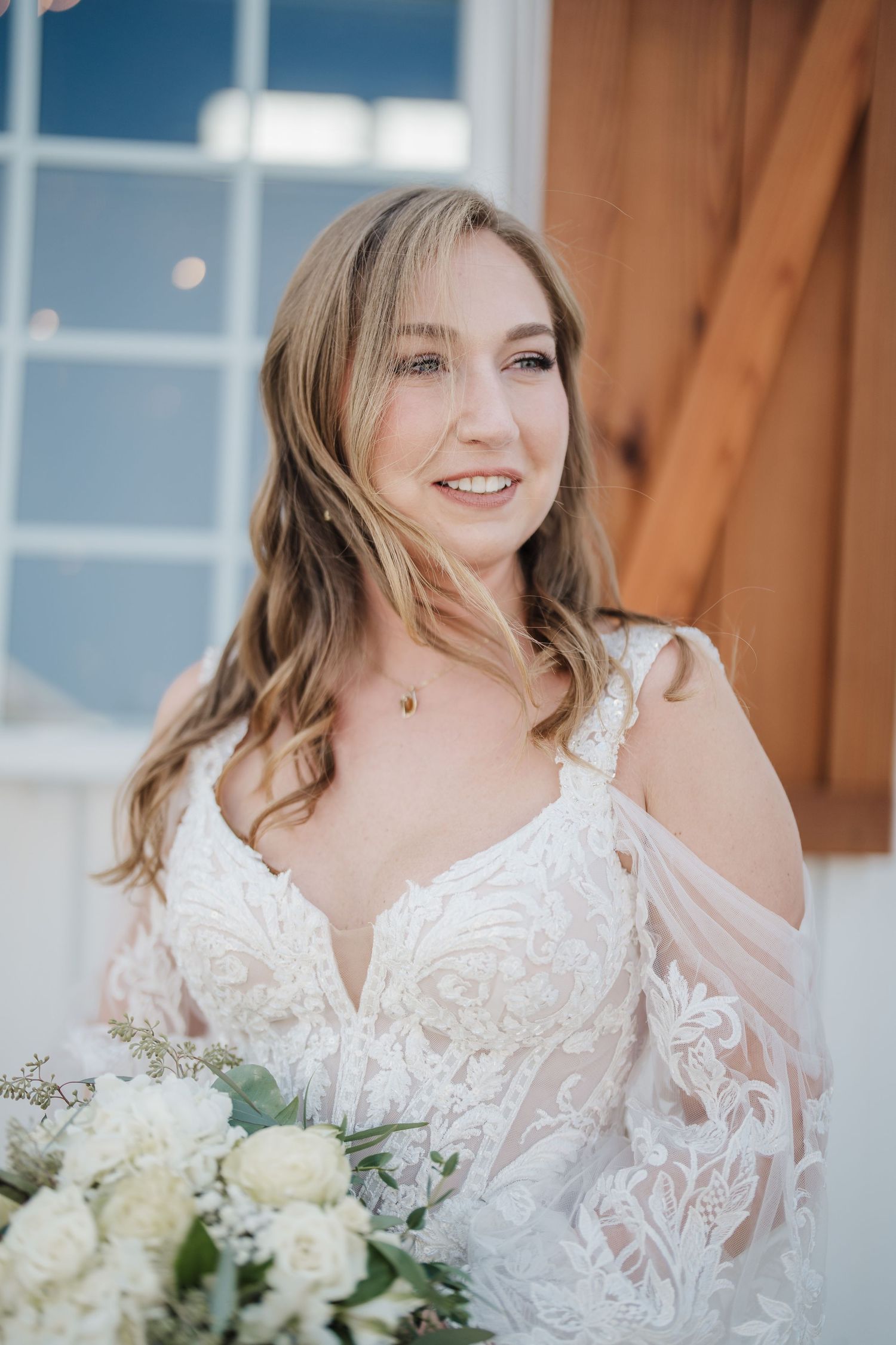 A bride in lace holds a white bouquet while posing against a wooden wall.