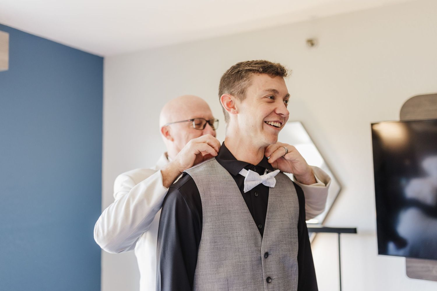 Two people share a lighthearted moment while adjusting formal wear in a blue-walled room.