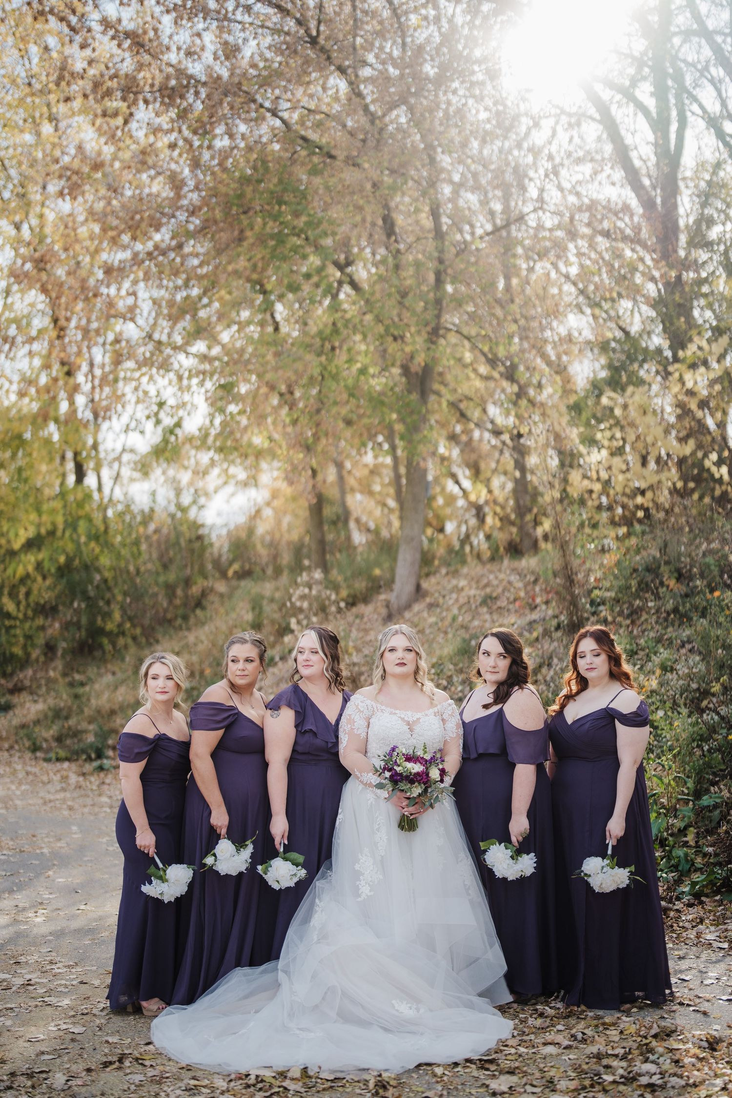 Bridal party in purple dresses poses with bride on a fall day with autumn foliage in background.