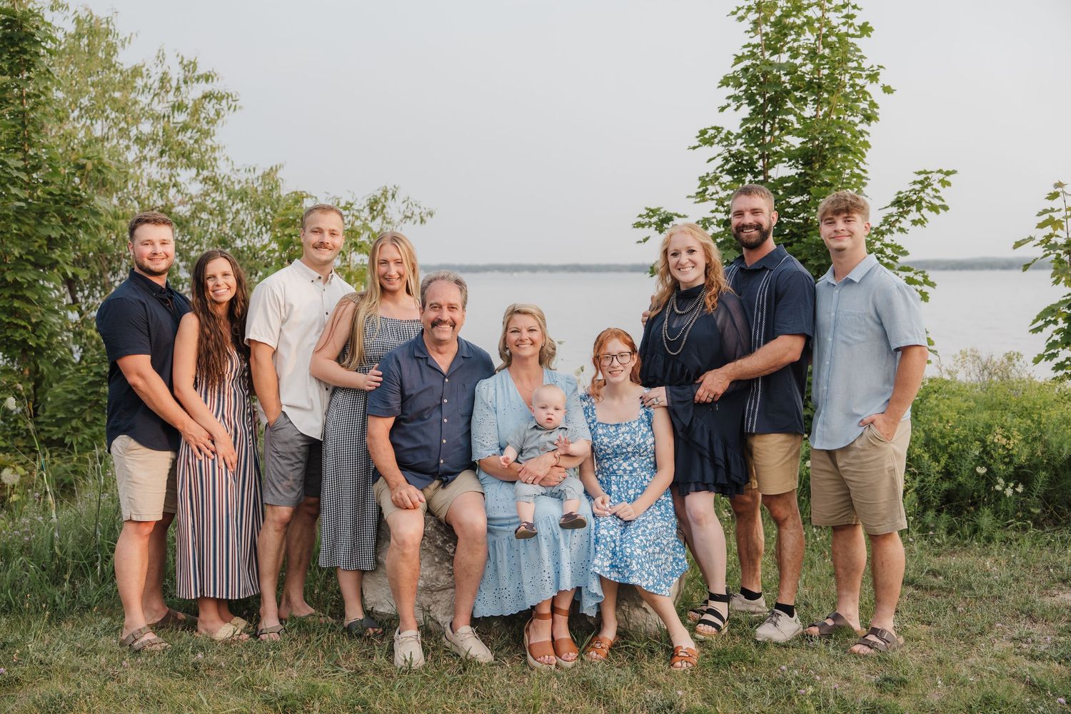 A large family of nine poses together outdoors near a lake, dressed in casual summer attire, smiling warmly.