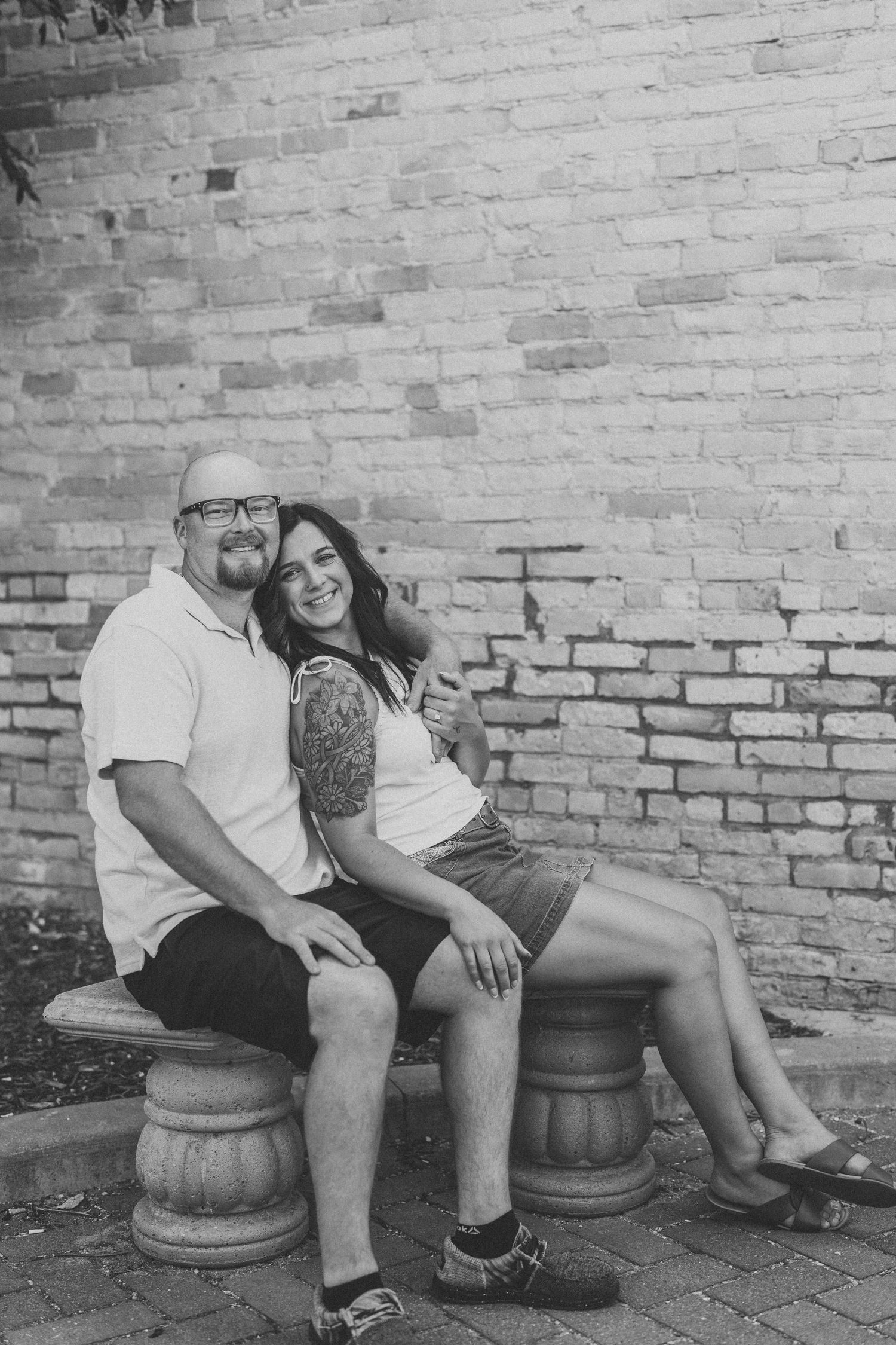Black and white photo of a smiling couple sitting on a stone bench against a brick wall.