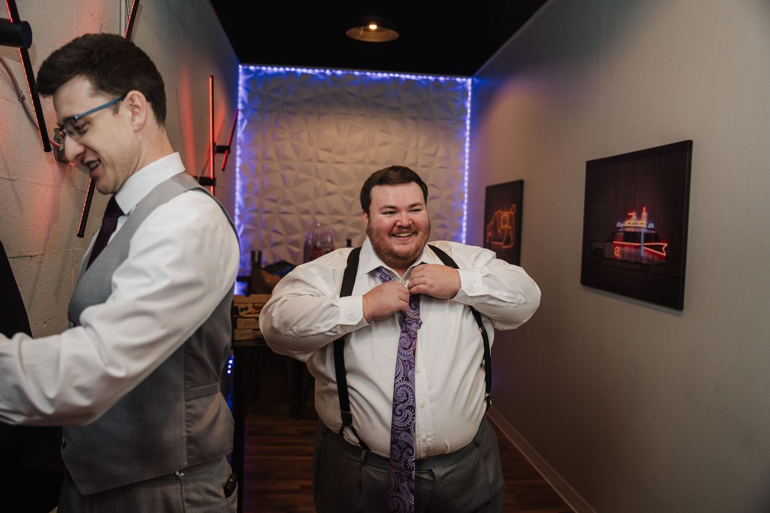 Groomsmen in white shirts and suspenders adjusting their attire in a hallway.