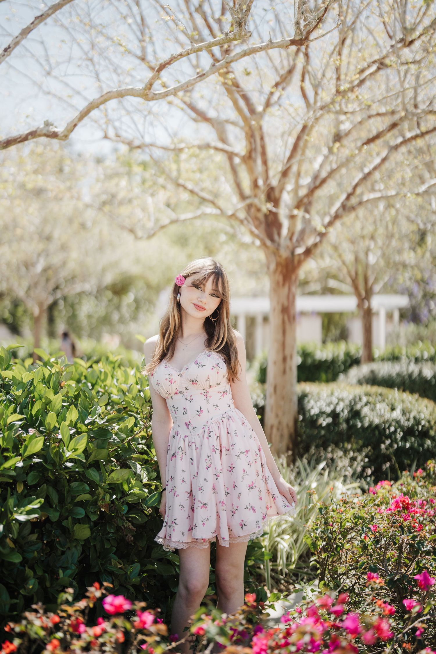 A floral spring dress twirls among blooming pink flowers and green bushes in a sunlit garden setting.