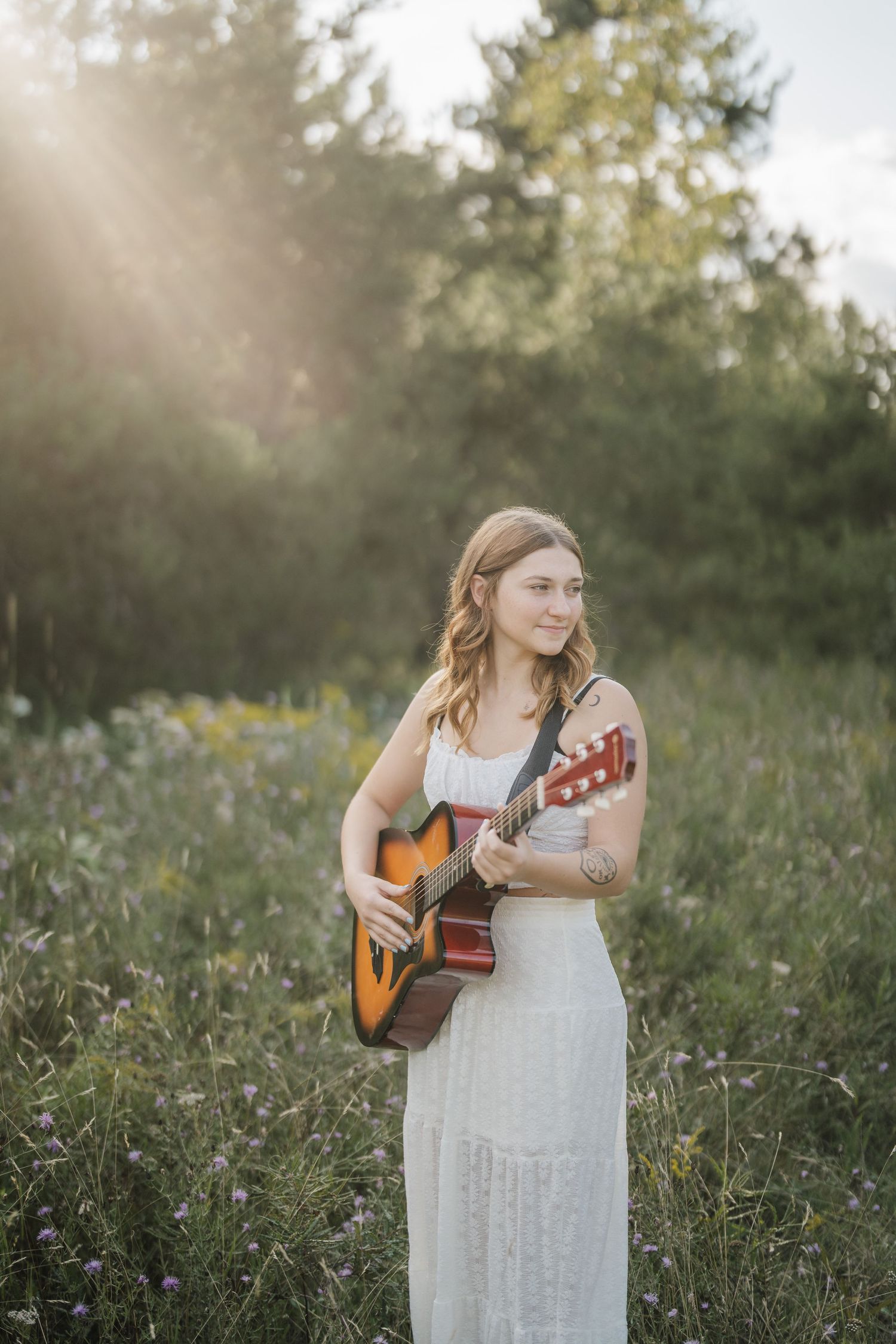 Musical portrait session in a sunlit meadow featuring a white sundress and acoustic guitar.