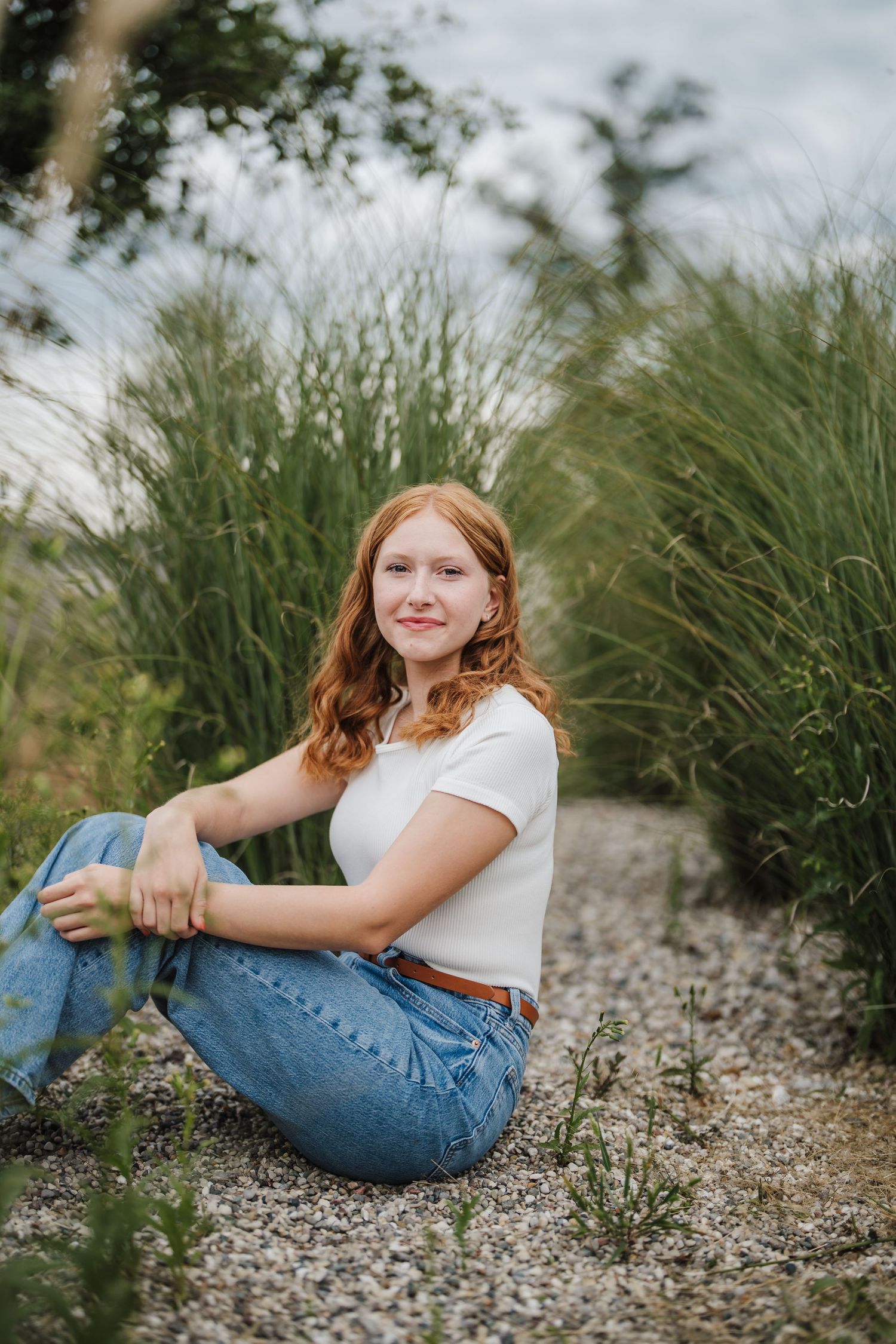 A person in a white crop top and jeans sits casually on rocky ground with tall ornamental grasses in the background.