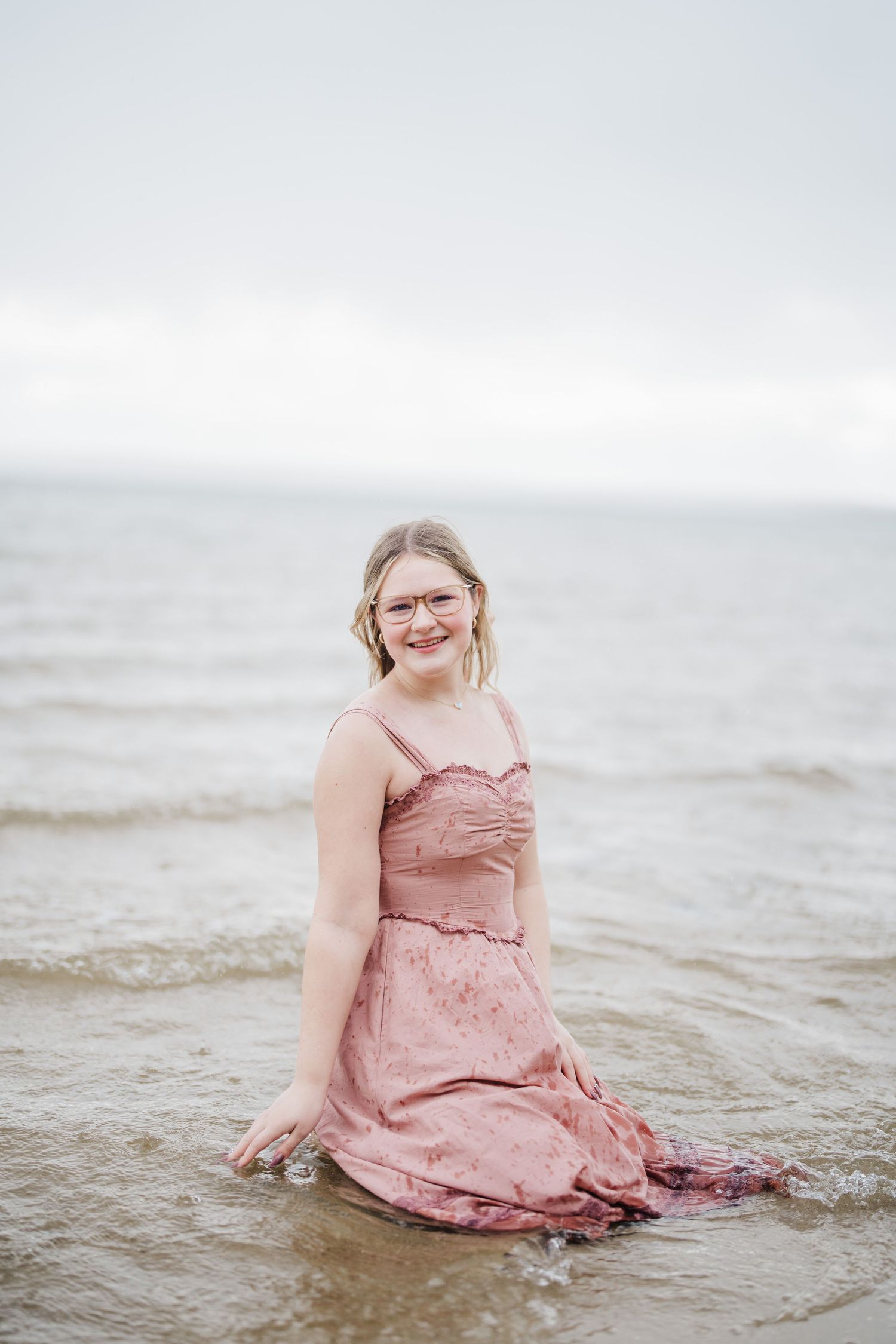A person in a pink dress sits joyfully in shallow ocean water during a beach photoshoot.