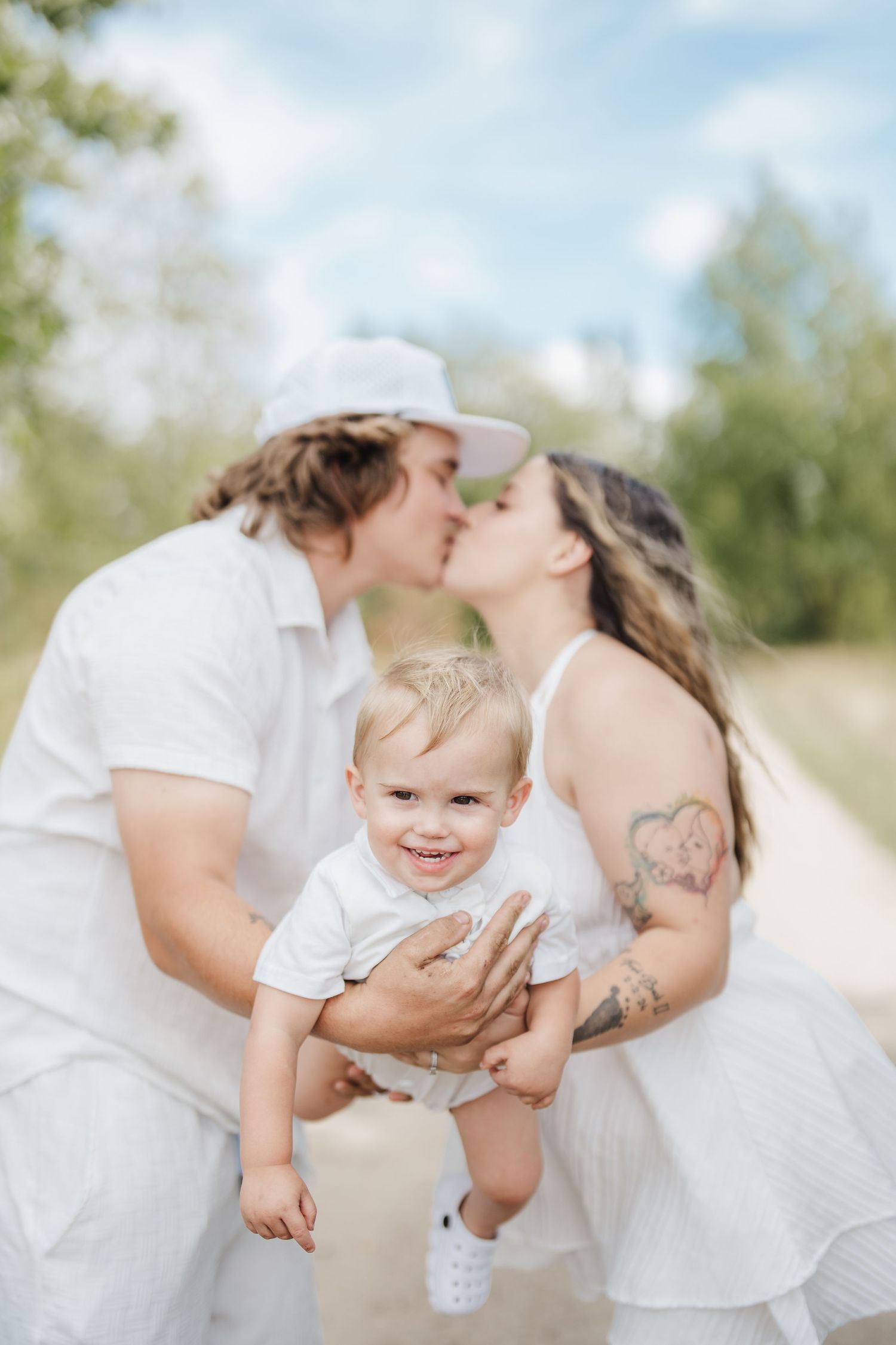 Family in white outfits shares tender moments on a beach with blurred natural background.