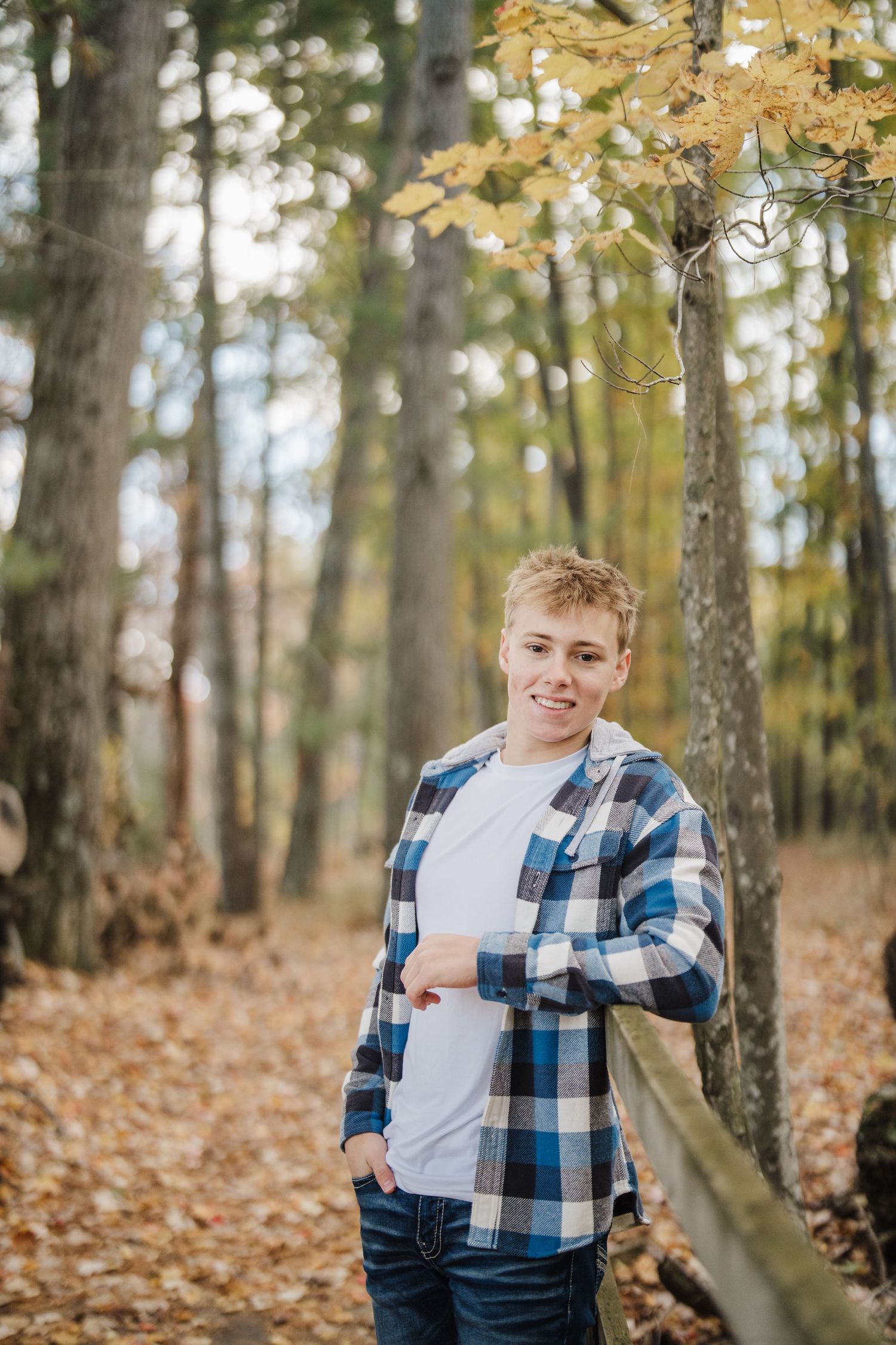 Student poses casually in autumn forest wearing blue plaid flannel shirt and jeans.