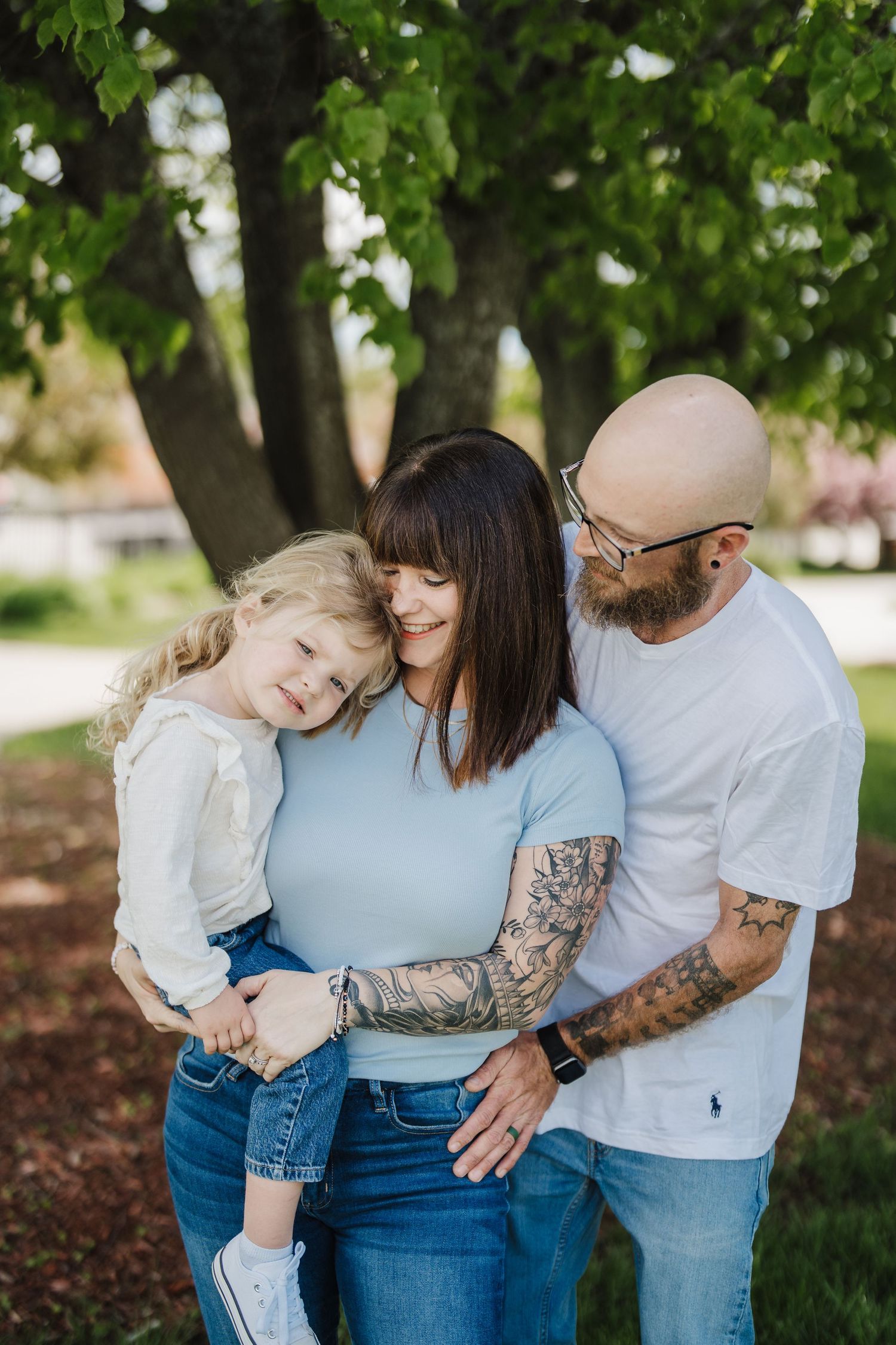 A family shares tender moments together in a park setting with green trees and natural lighting.