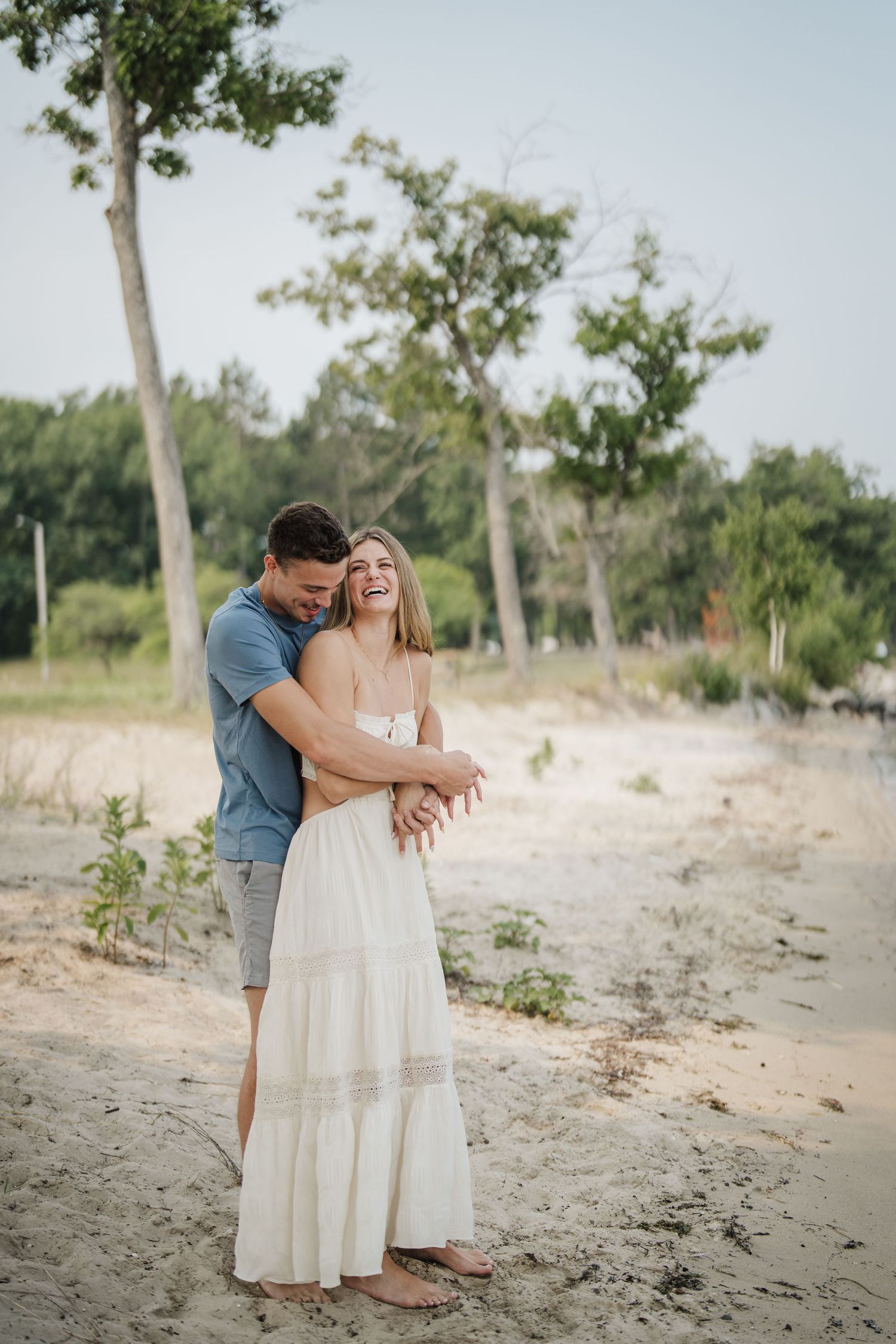 Couple shares tender moments on a sandy beach with trees and greenery in the background.