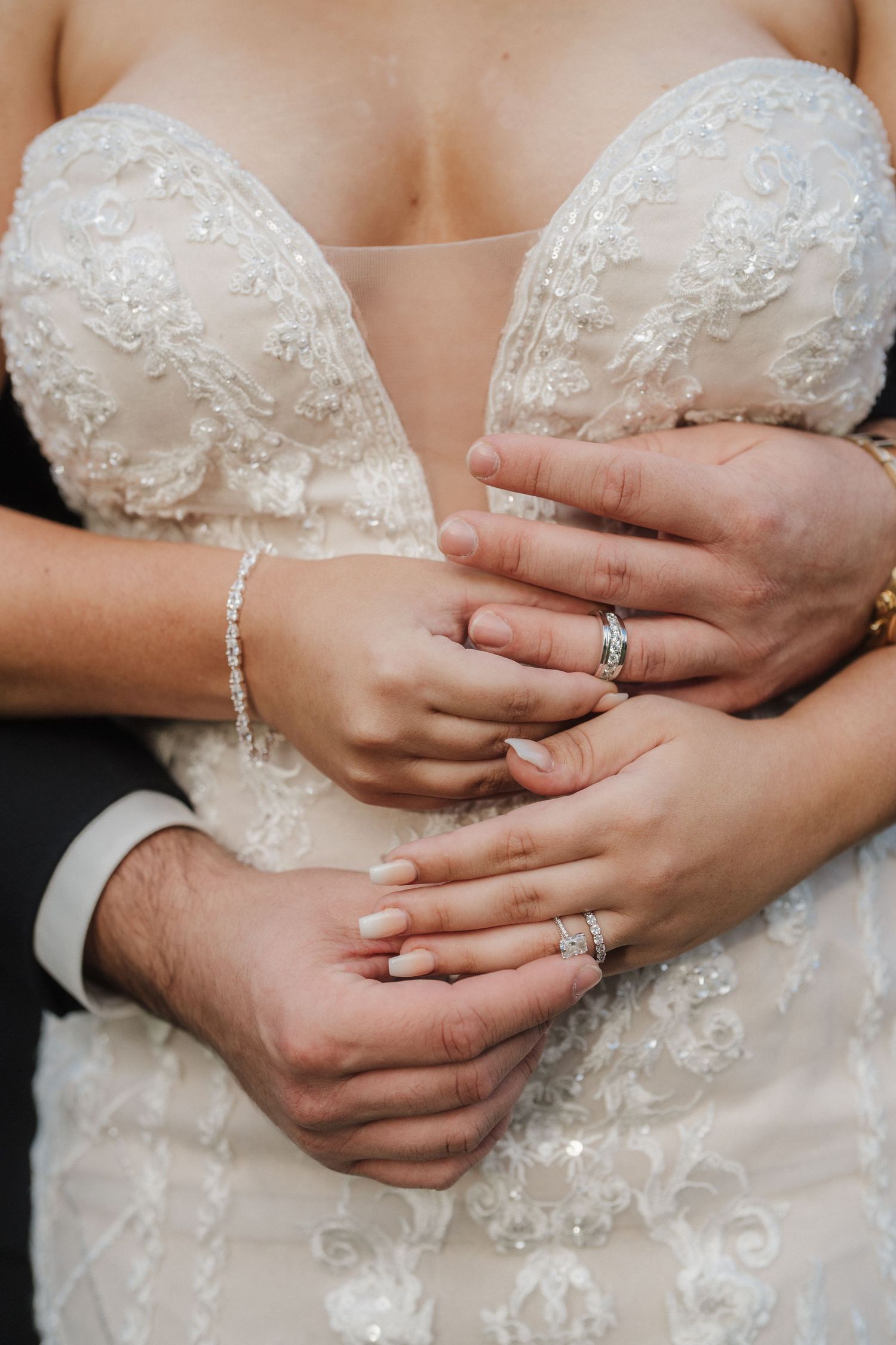 Close-up of hands wearing wedding rings against lace wedding dress detail.