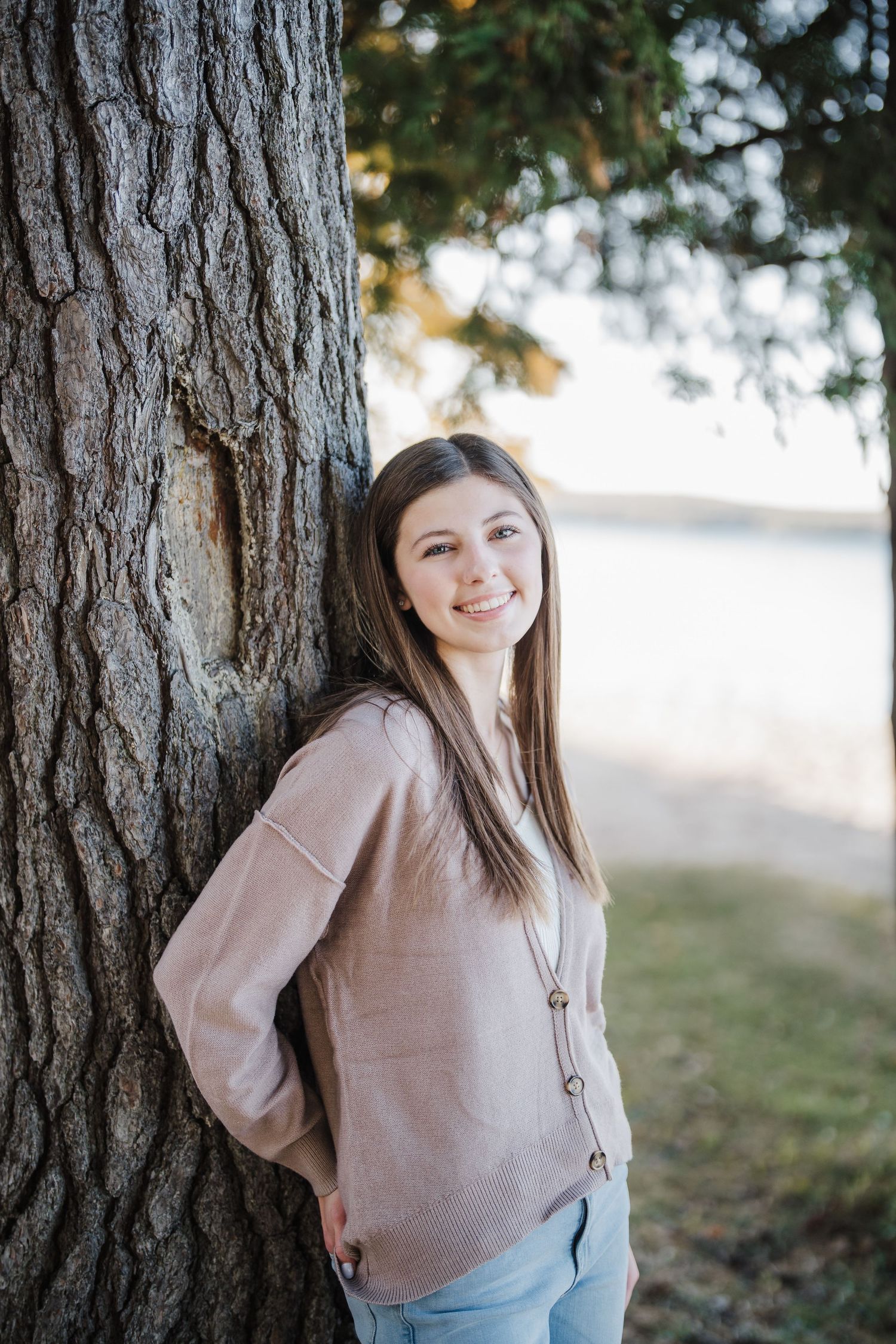 A person in a beige sweater poses against a tree trunk in an outdoor portrait setting.