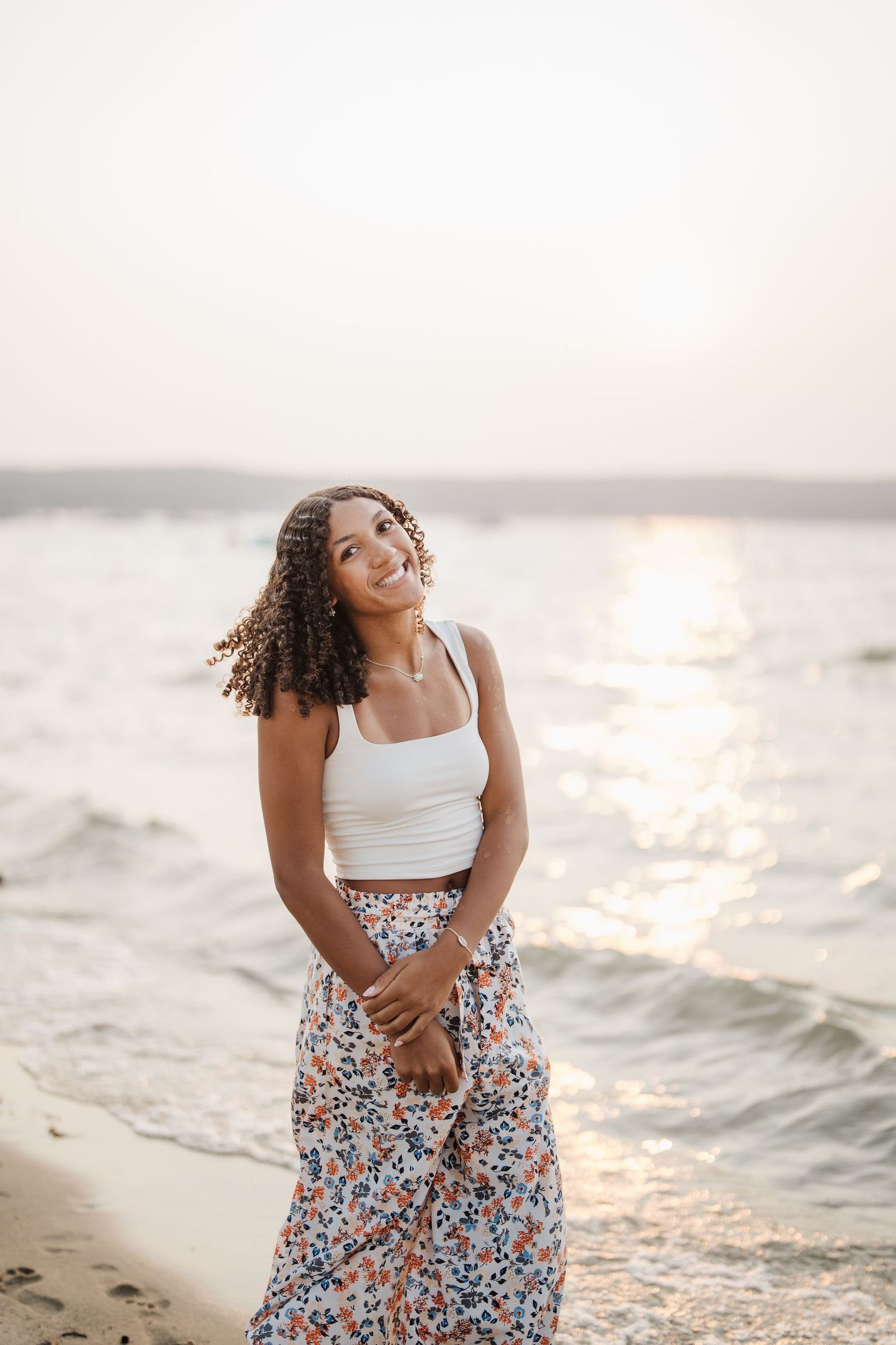A beachside portrait at sunset wearing a white tank top and floral skirt with waves in the background.
