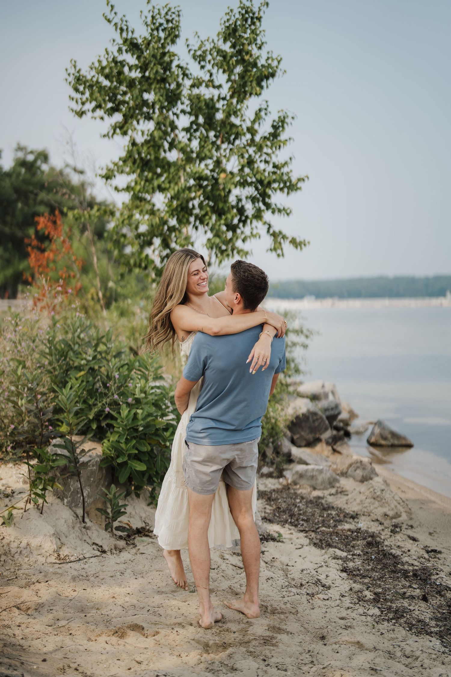 A couple embraces on a scenic rocky beach shoreline with trees and water in the background.