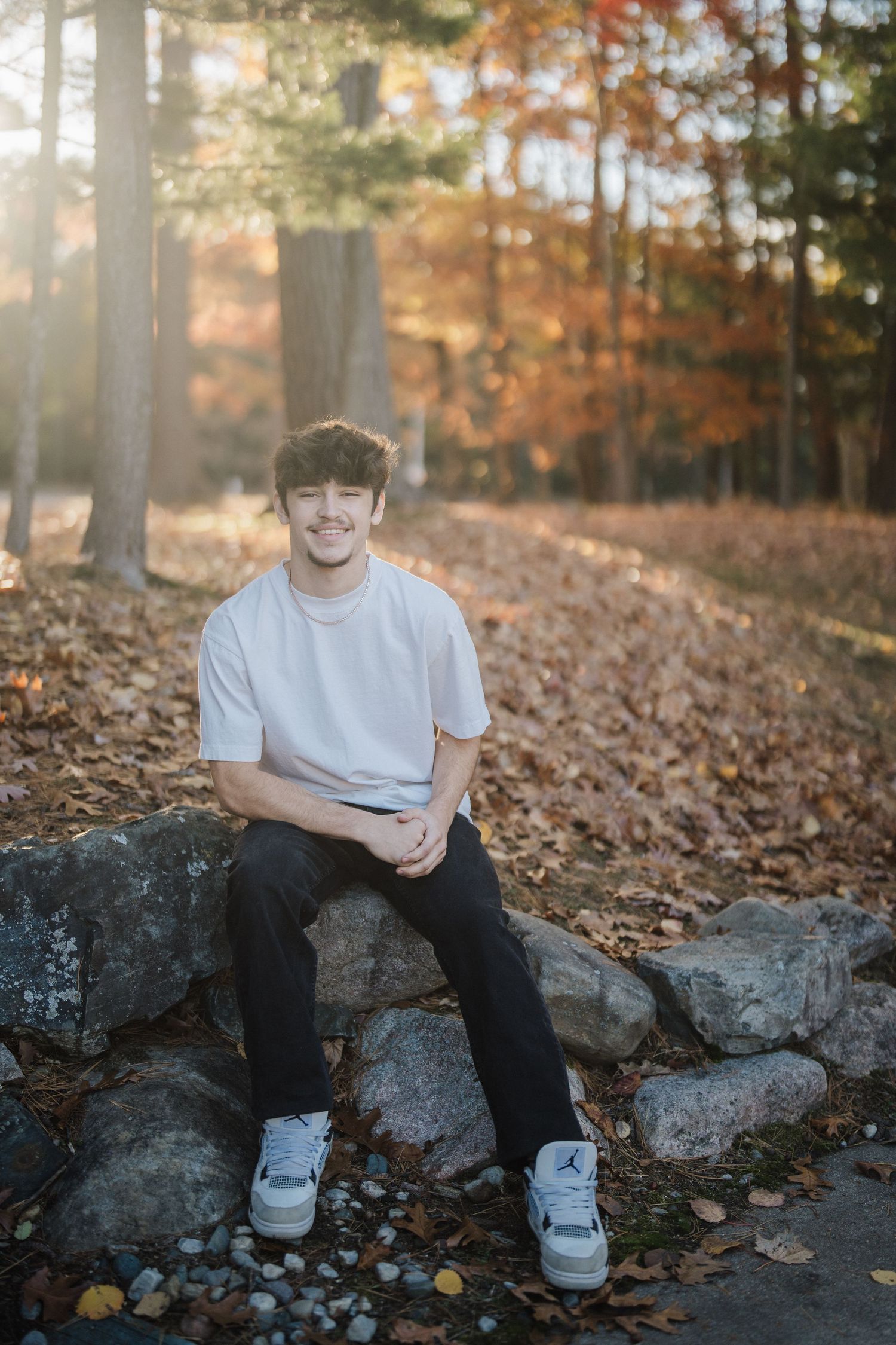 A person sits on large rocks surrounded by fallen autumn leaves in a forest setting wearing casual attire.