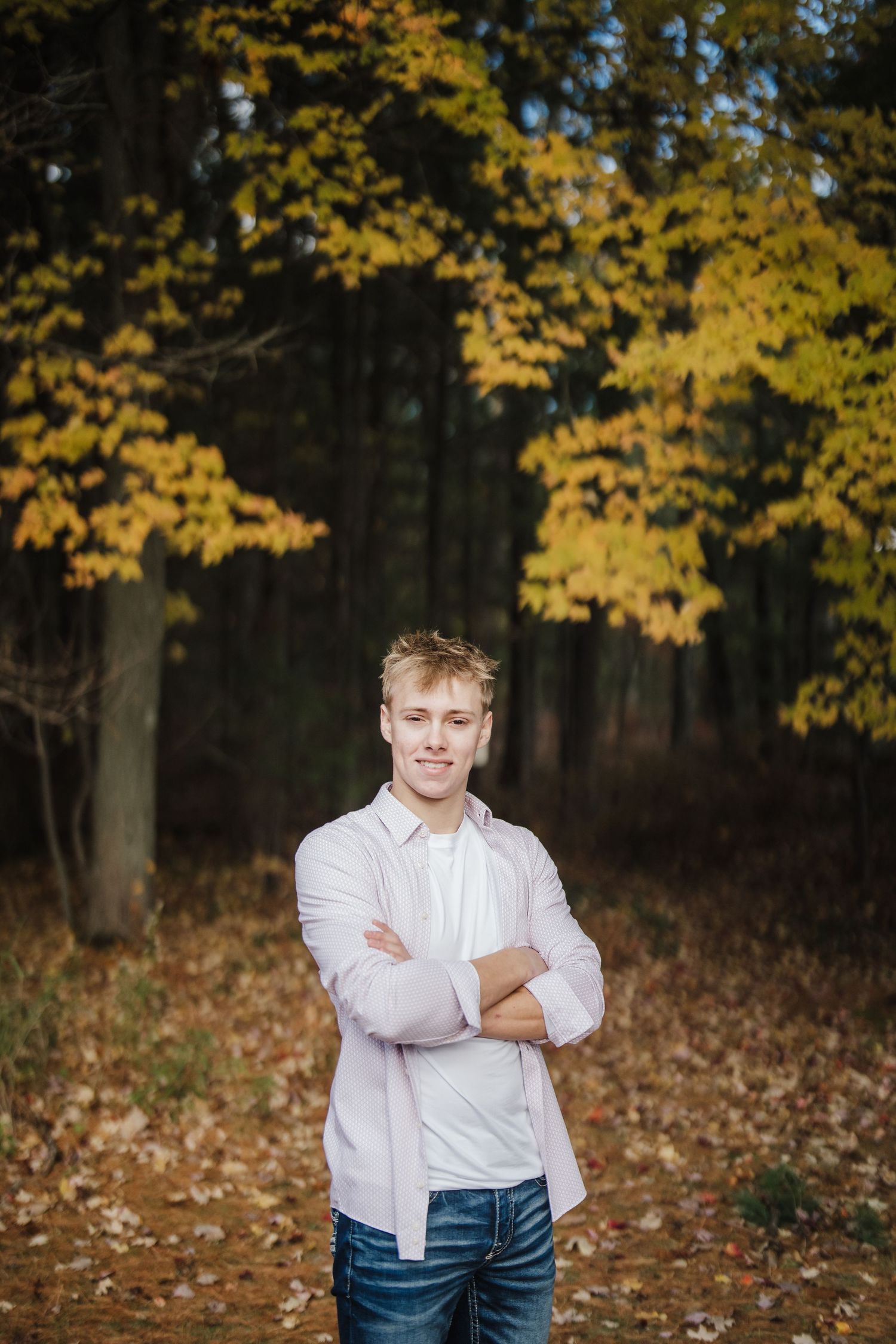 Person in white button-down shirt and jeans smiles warmly against fall foliage background.