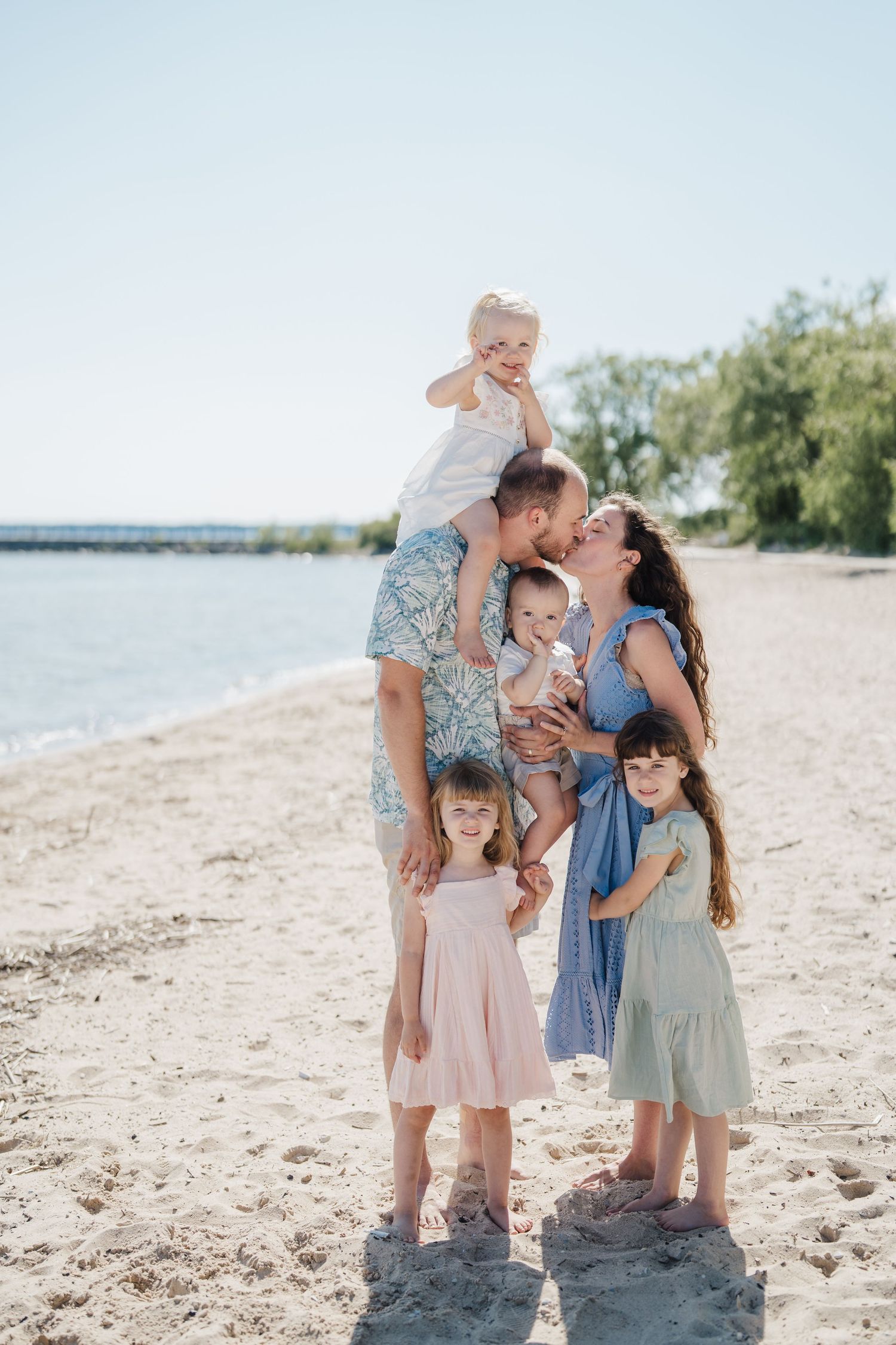 Family enjoying a sunny day together on a sandy beach with trees and water in the background.