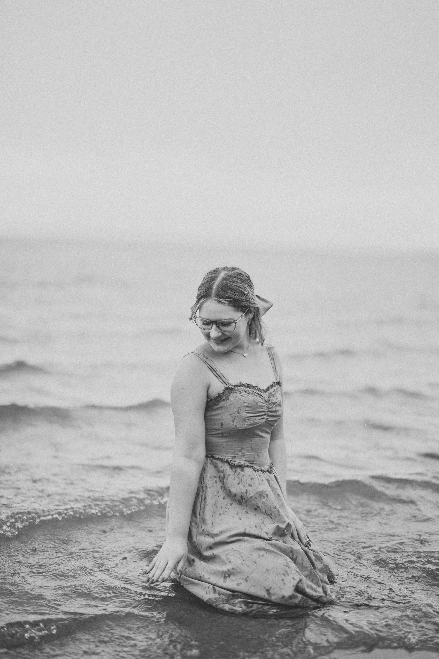 A person in a floral dress sits gracefully in shallow ocean waters, creating a dreamy black and white beach scene.