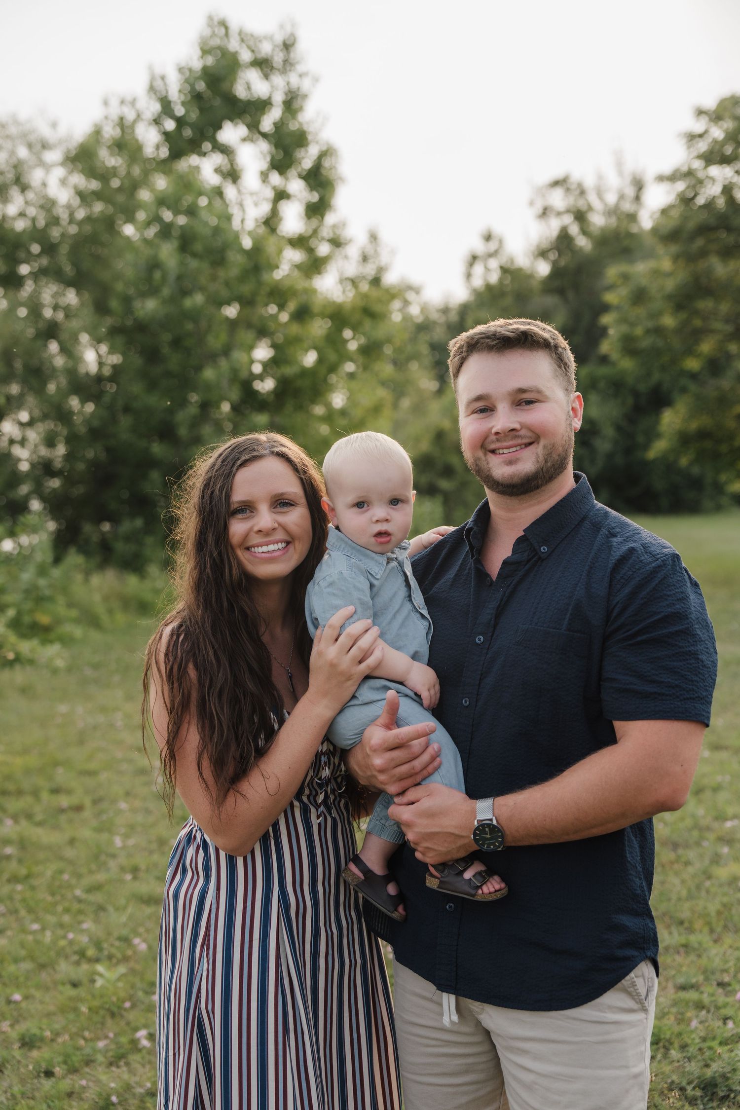 A smiling young family with a baby boy poses outdoors in a green field for a portrait photo.