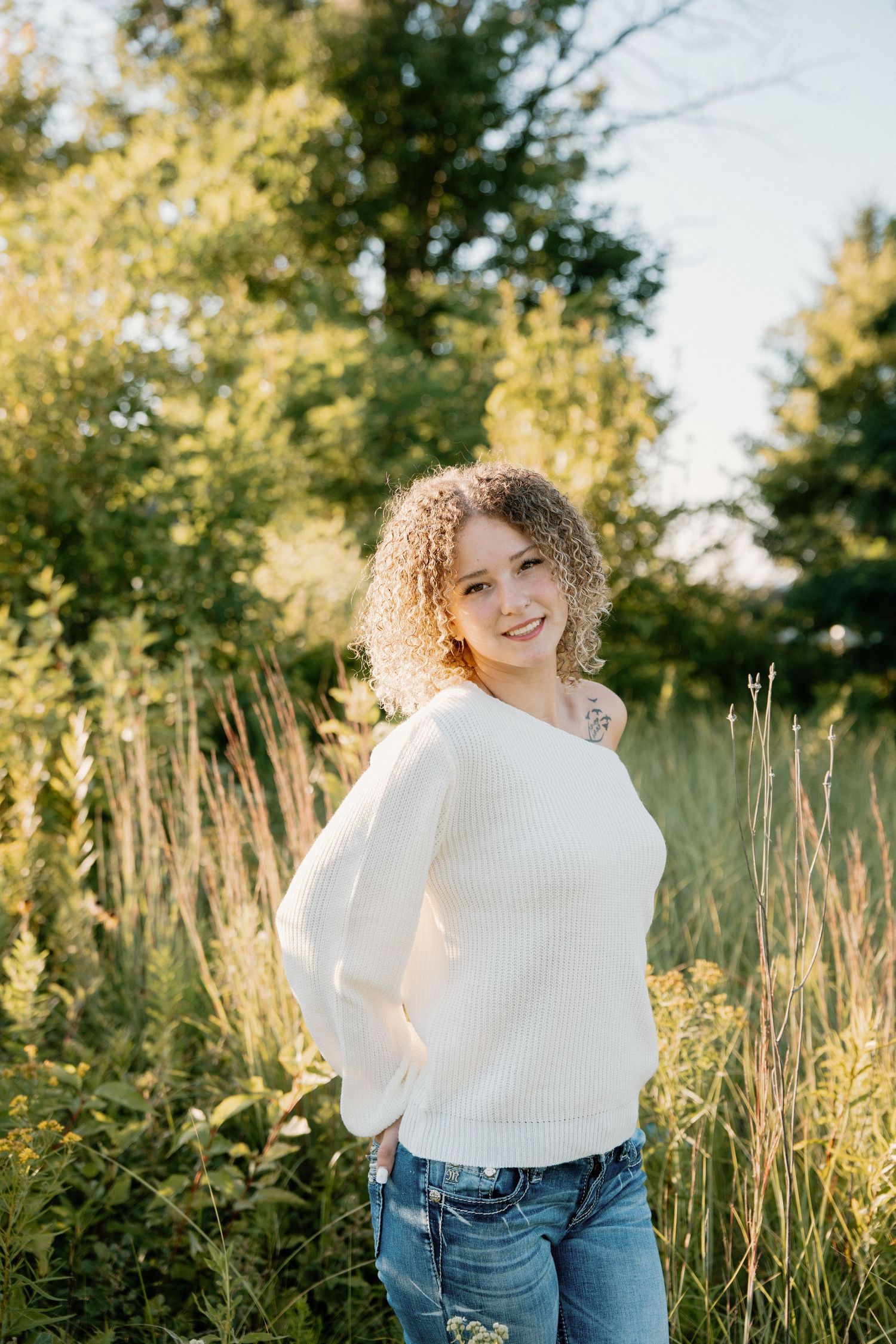 Smiling young woman with curly hair in white sweater and jeans posing outdoors in sunlit green field.