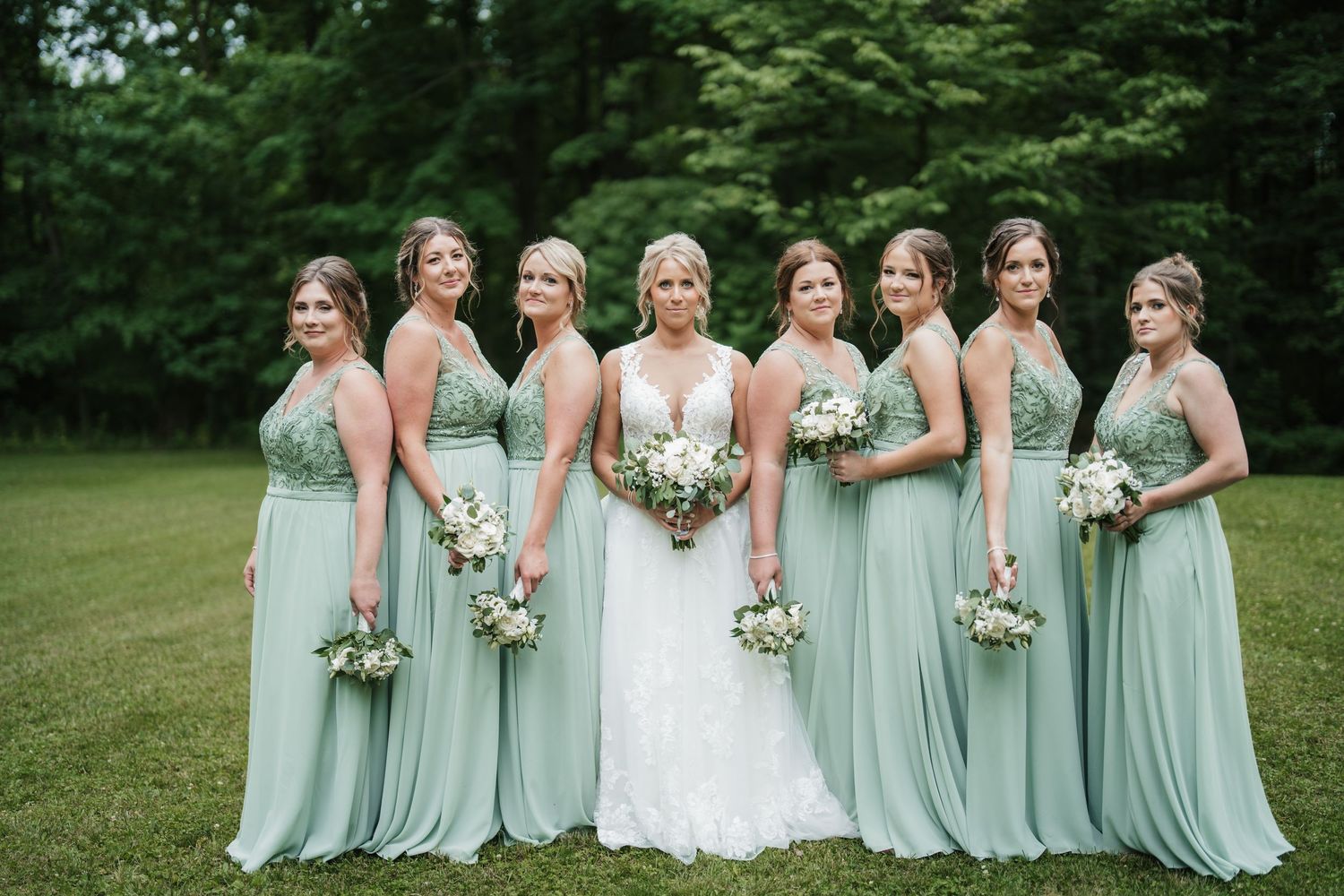 Bridal party in mint green dresses poses on lawn with white bouquets.