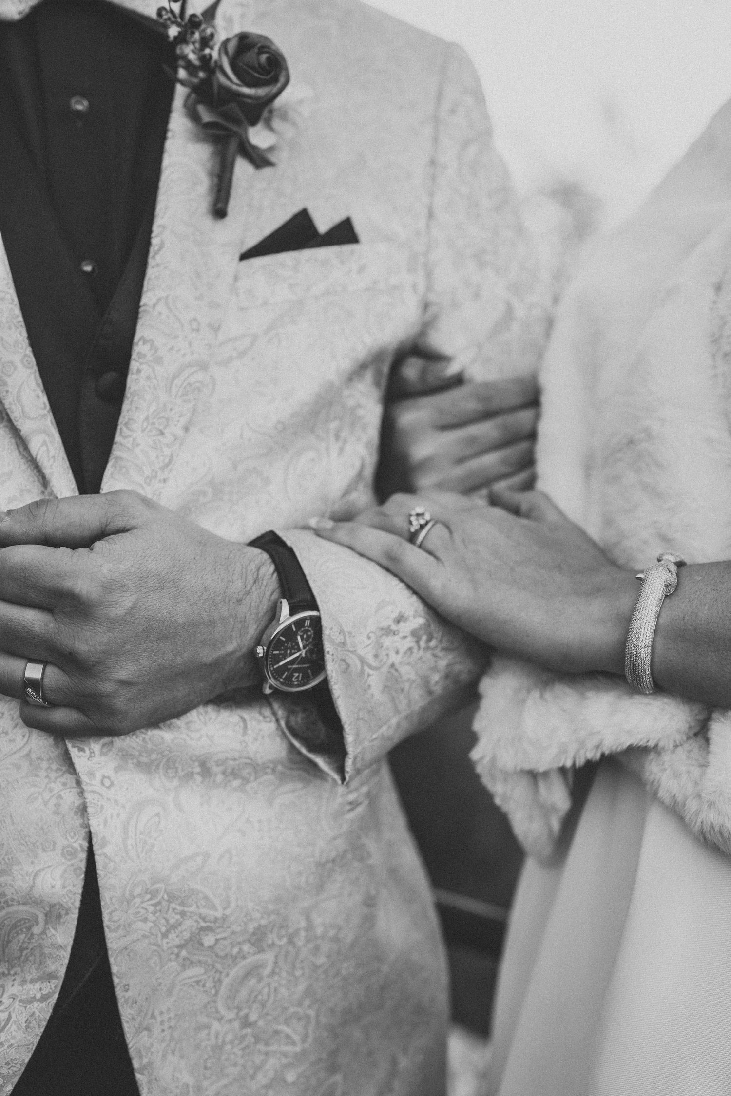 Close-up of wedding couple's hands showing rings and formal attire details in black and white.