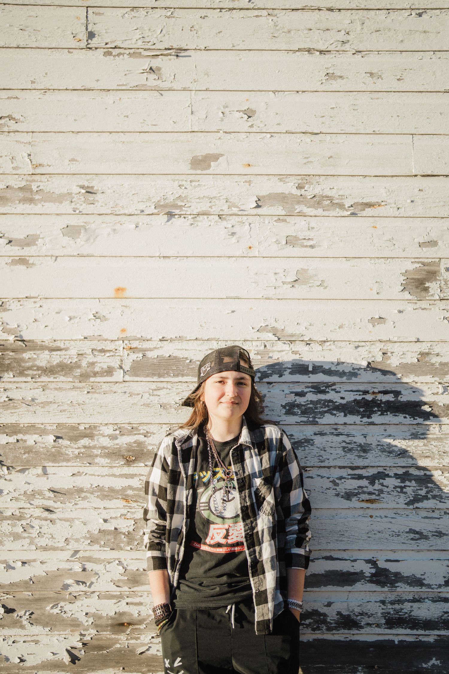 A casual outdoor portrait against a weathered white wooden wall wearing plaid and camouflage.