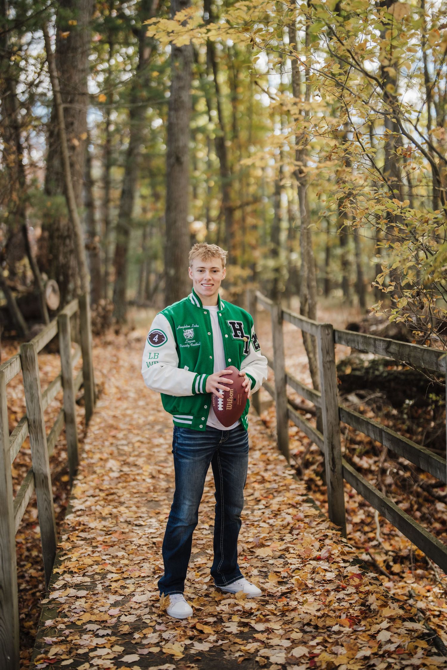 Person wearing green varsity jacket holds football while walking through leaf-covered forest path.