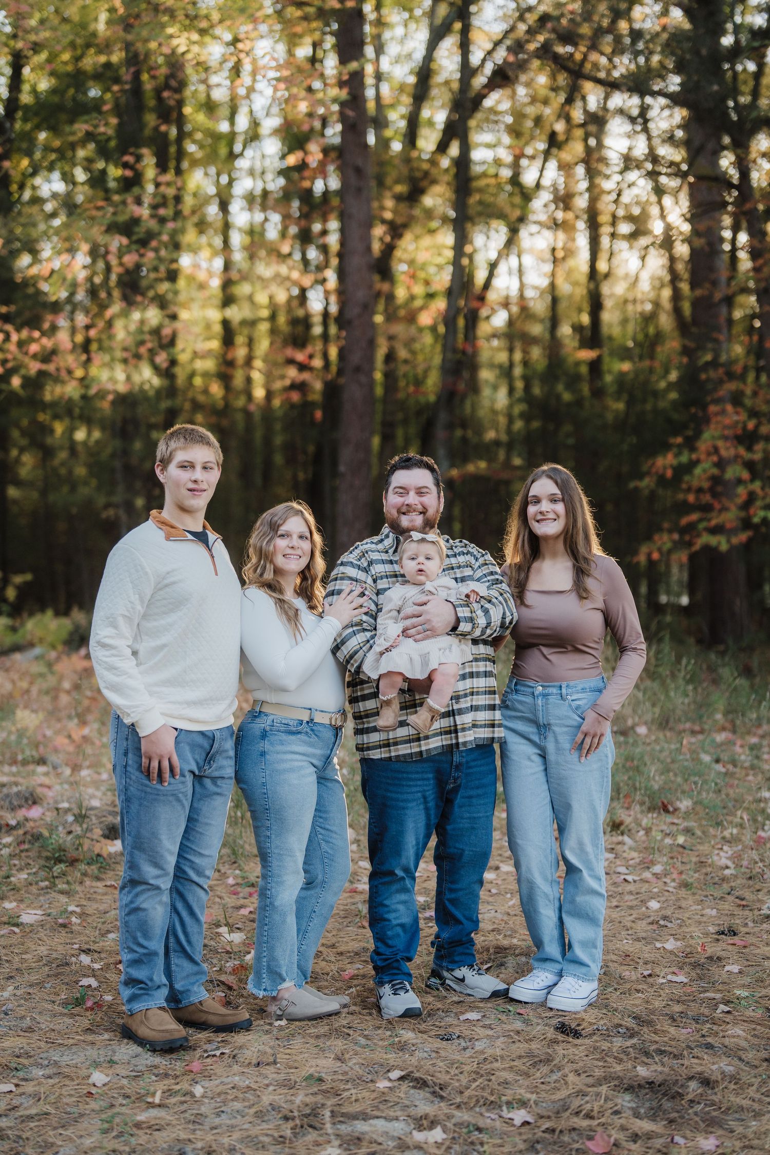 A group poses outdoors in casual attire sitting together on fallen autumn leaves in a wooded setting.