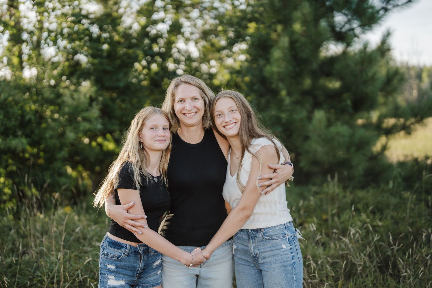 Three people in casual clothing pose together outdoors against a natural green background in warm evening light.