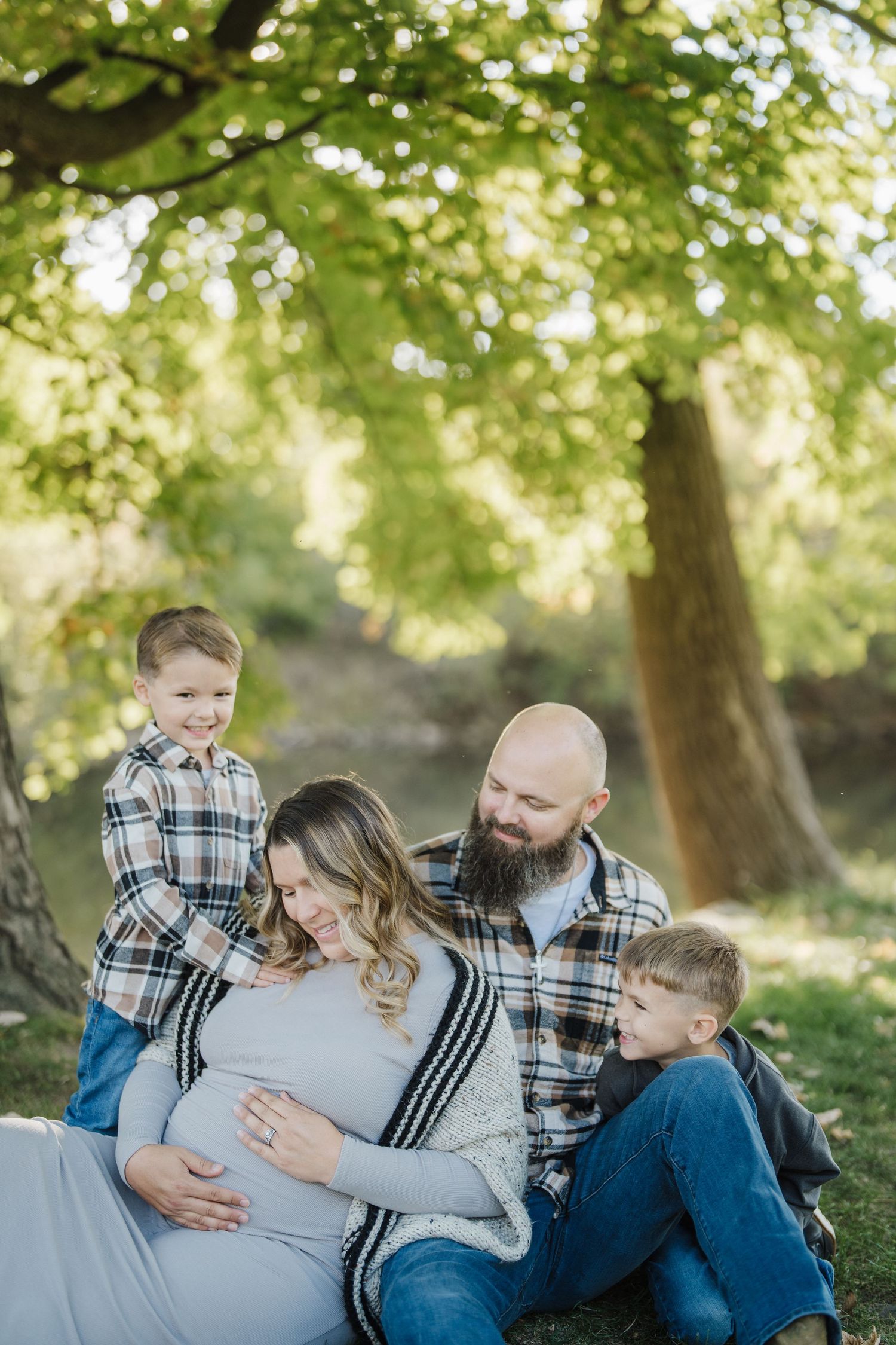 A family shares joyful moments together while sitting outdoors in a sunlit park with green trees in the background.