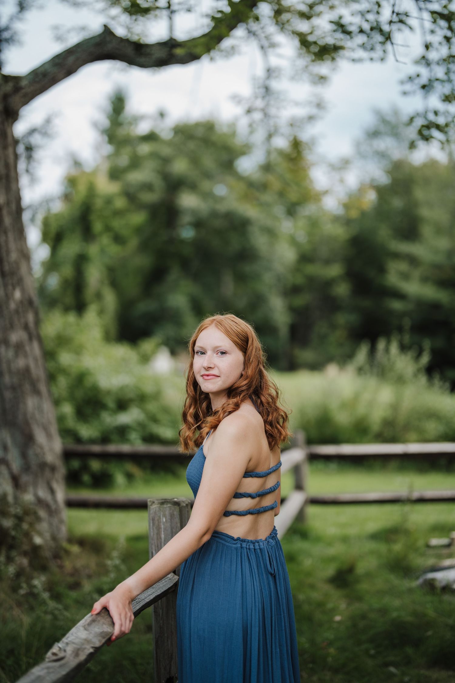 A person in a blue dress poses outdoors in a natural setting with trees and greenery in the background.