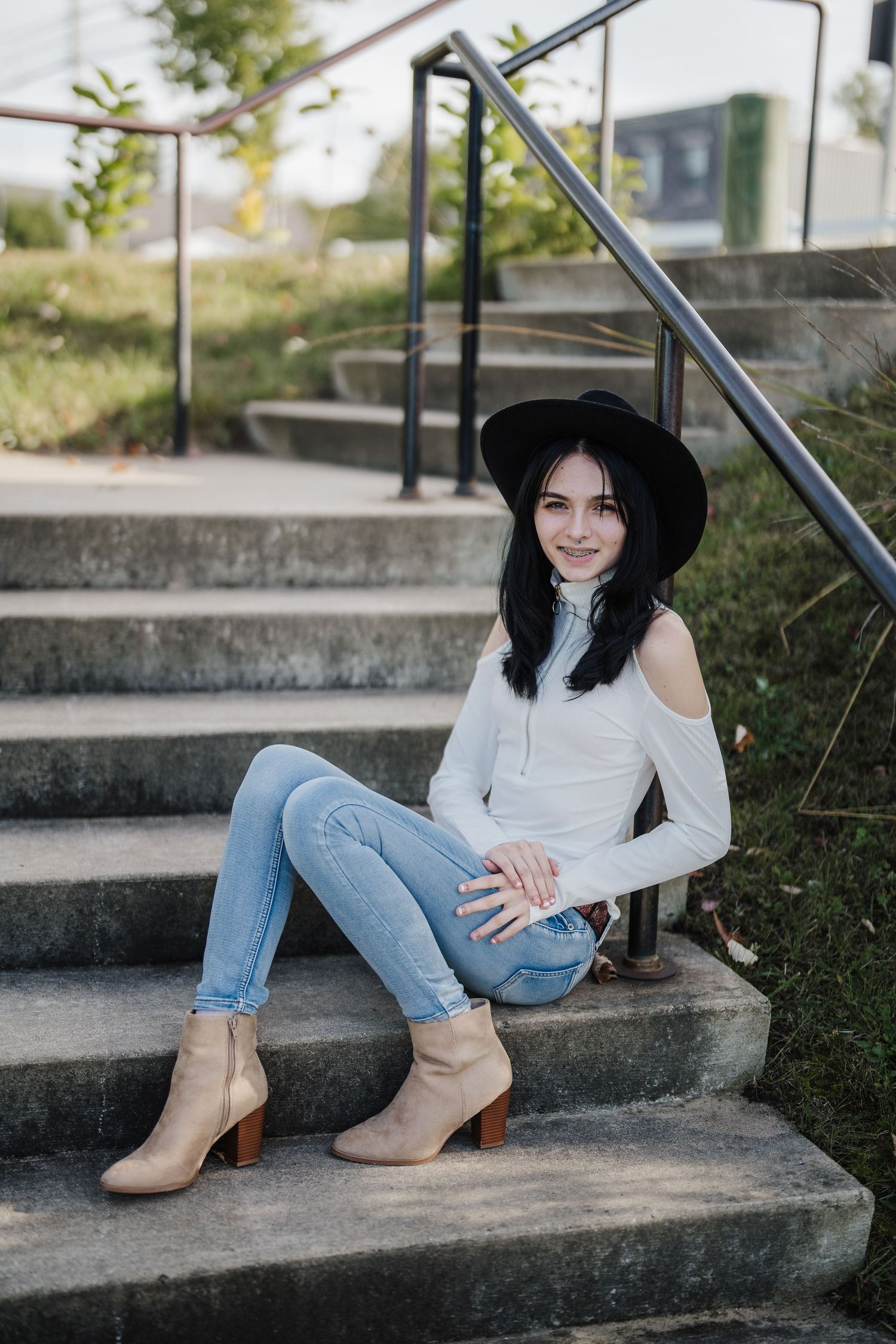 Person in white sweater, jeans and brown boots sits casually on concrete steps wearing a black hat.