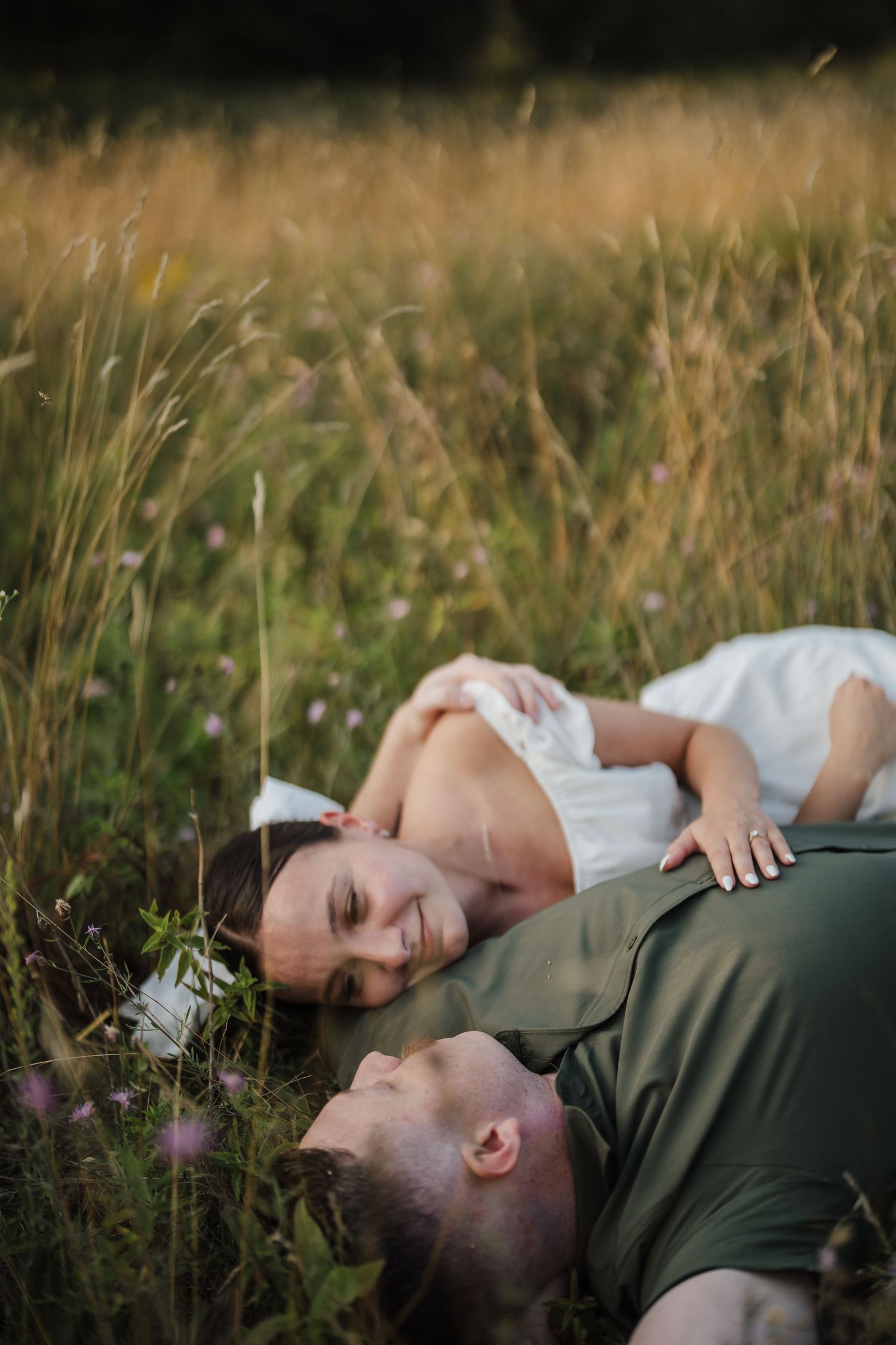 A couple lies together in tall grass during a romantic outdoor photo session at golden hour.