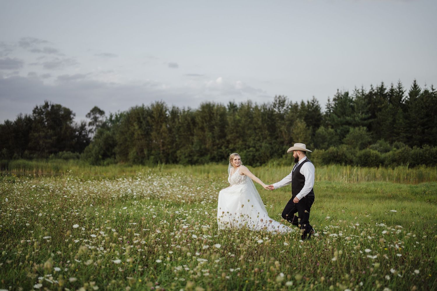 A couple in wedding attire walk through a wildflower meadow surrounded by trees at dusk.