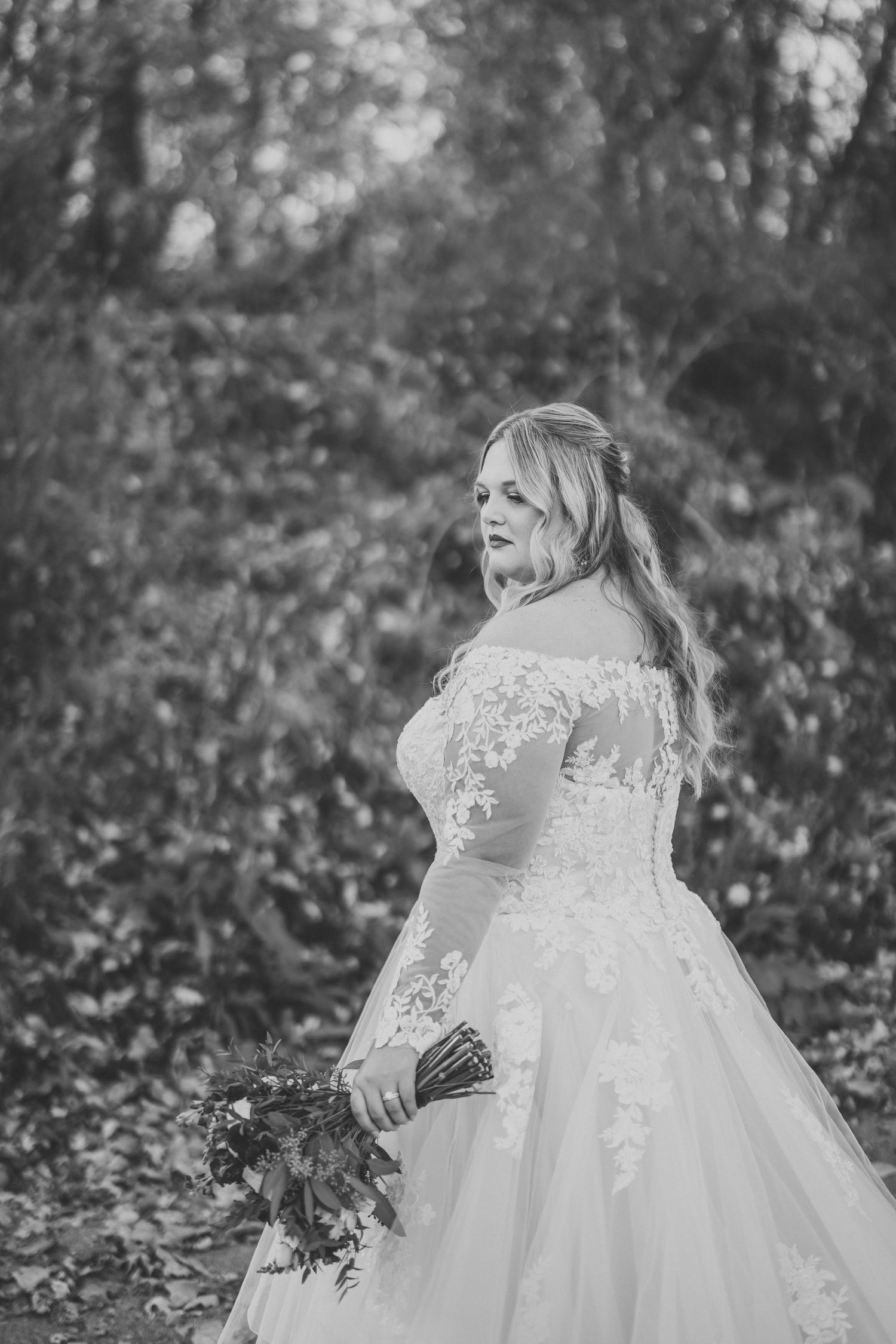 A black and white wedding portrait shows a bride in a lace gown holding a bouquet in a forest setting.