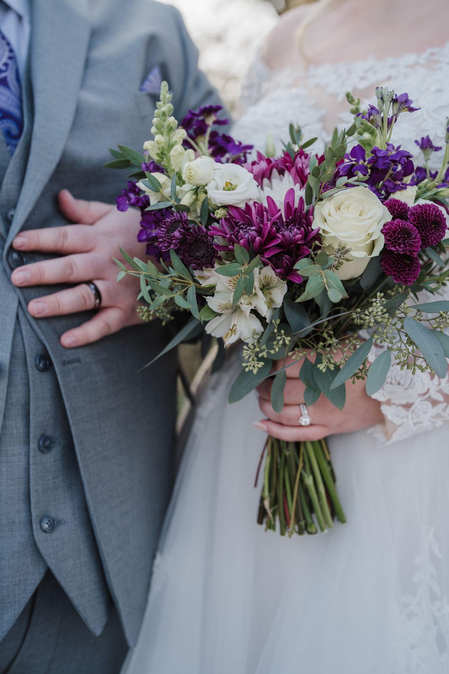 Close-up of wedding bouquet with purple and white flowers against grey suit.