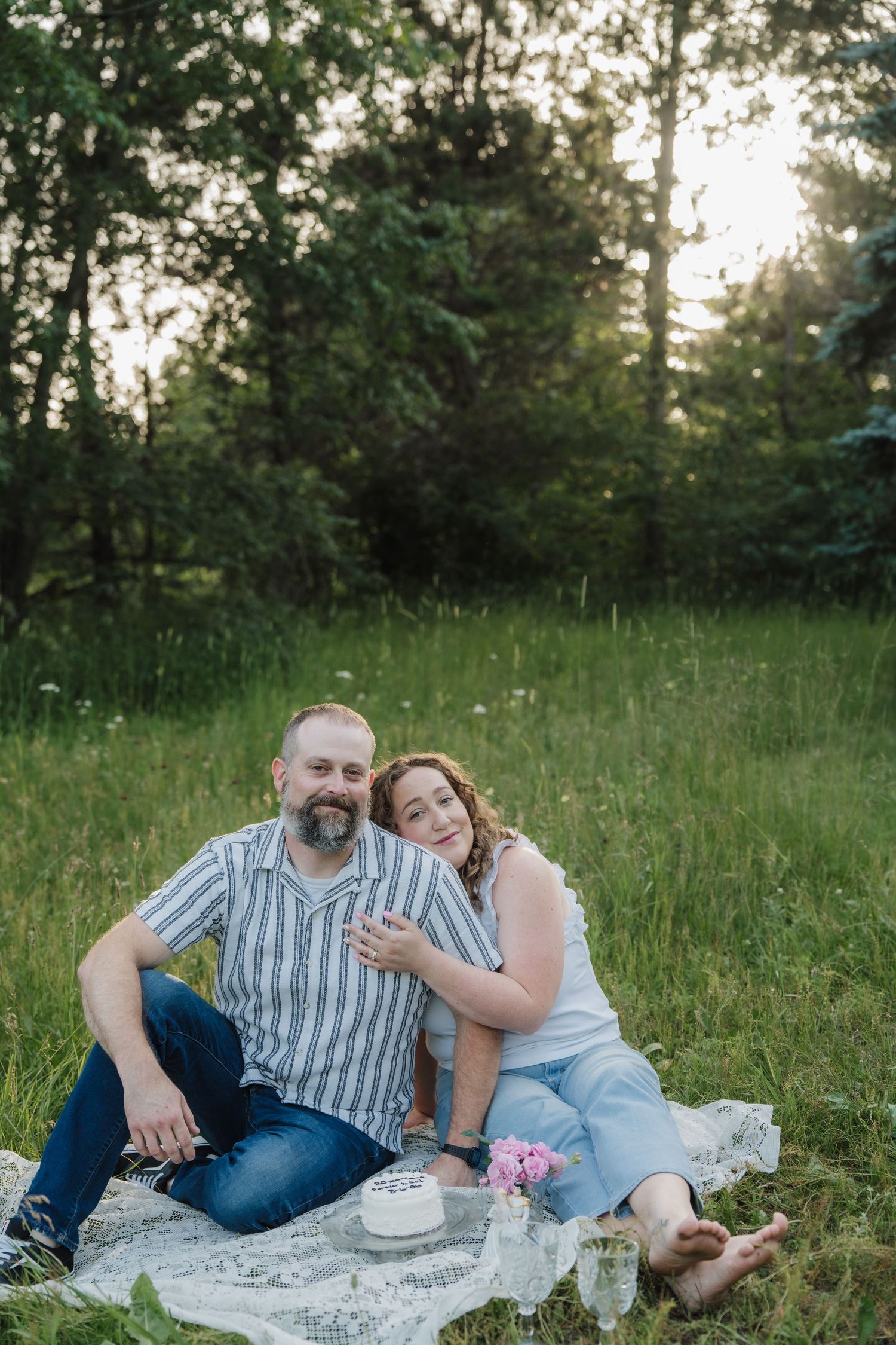 Couple sitting on a blanket in a green meadow, smiling and embracing during a romantic outdoor picnic at sunset.
