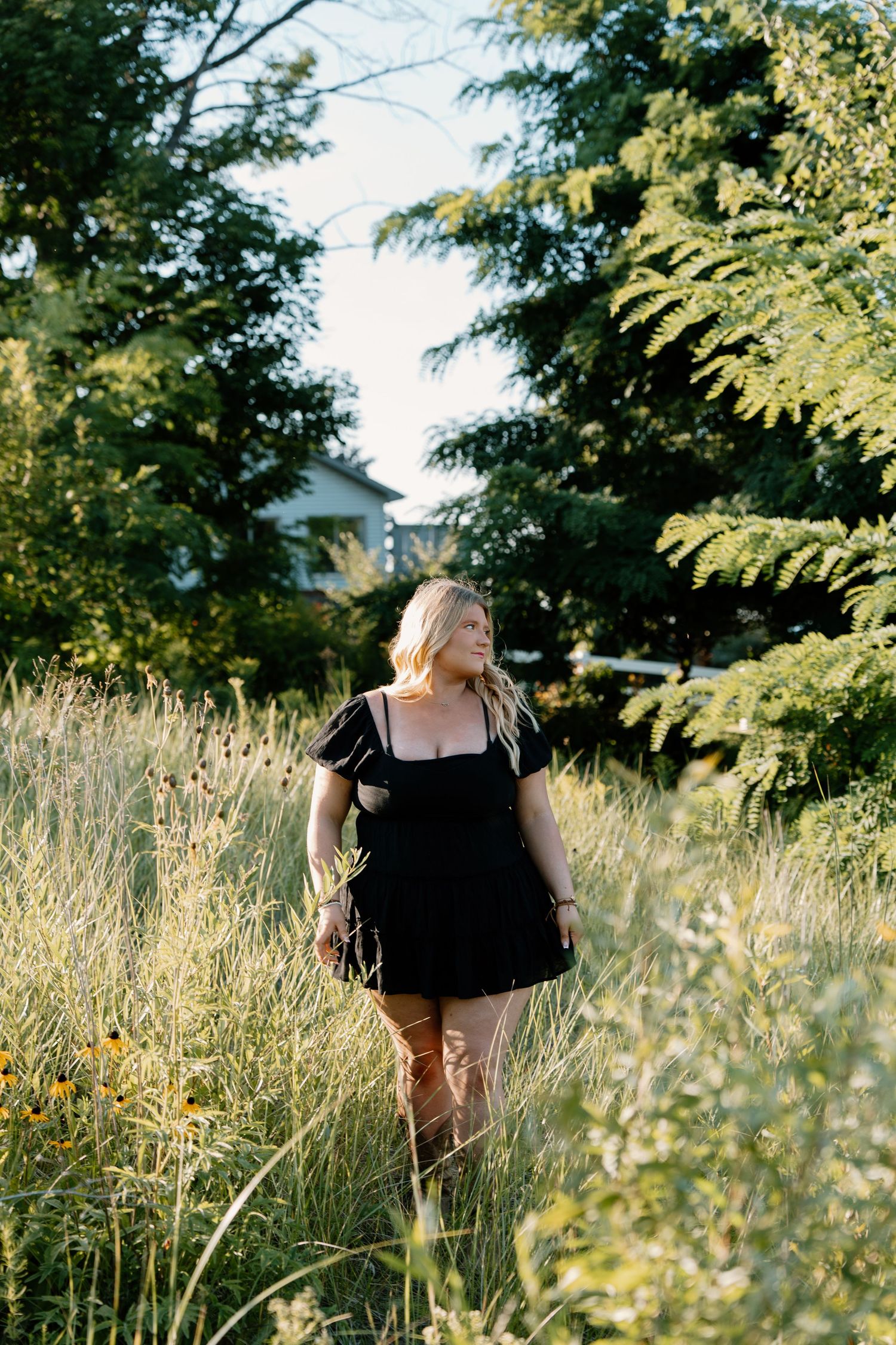 Woman in black mini dress standing in tall wildflower field surrounded by lush green trees on sunny day.