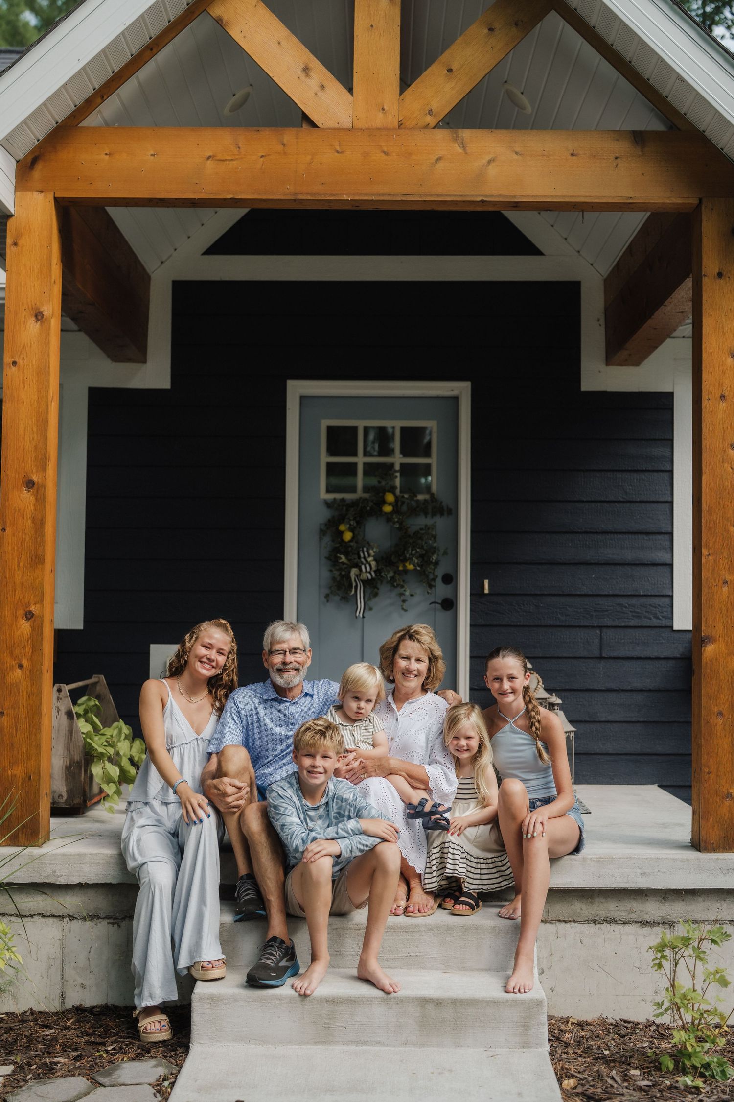 Multi-generational family of seven smiling together on porch steps of a dark blue cottage home.