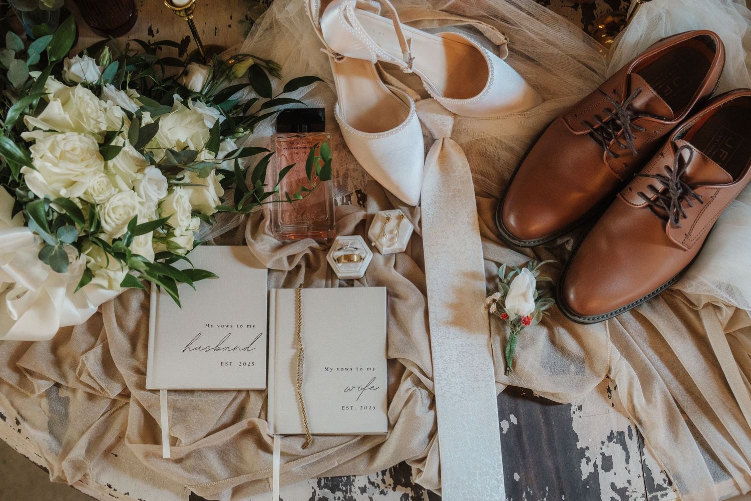 Wedding details flatlay with brown leather shoes, white floral bouquet, invitations and ribbons on neutral background.