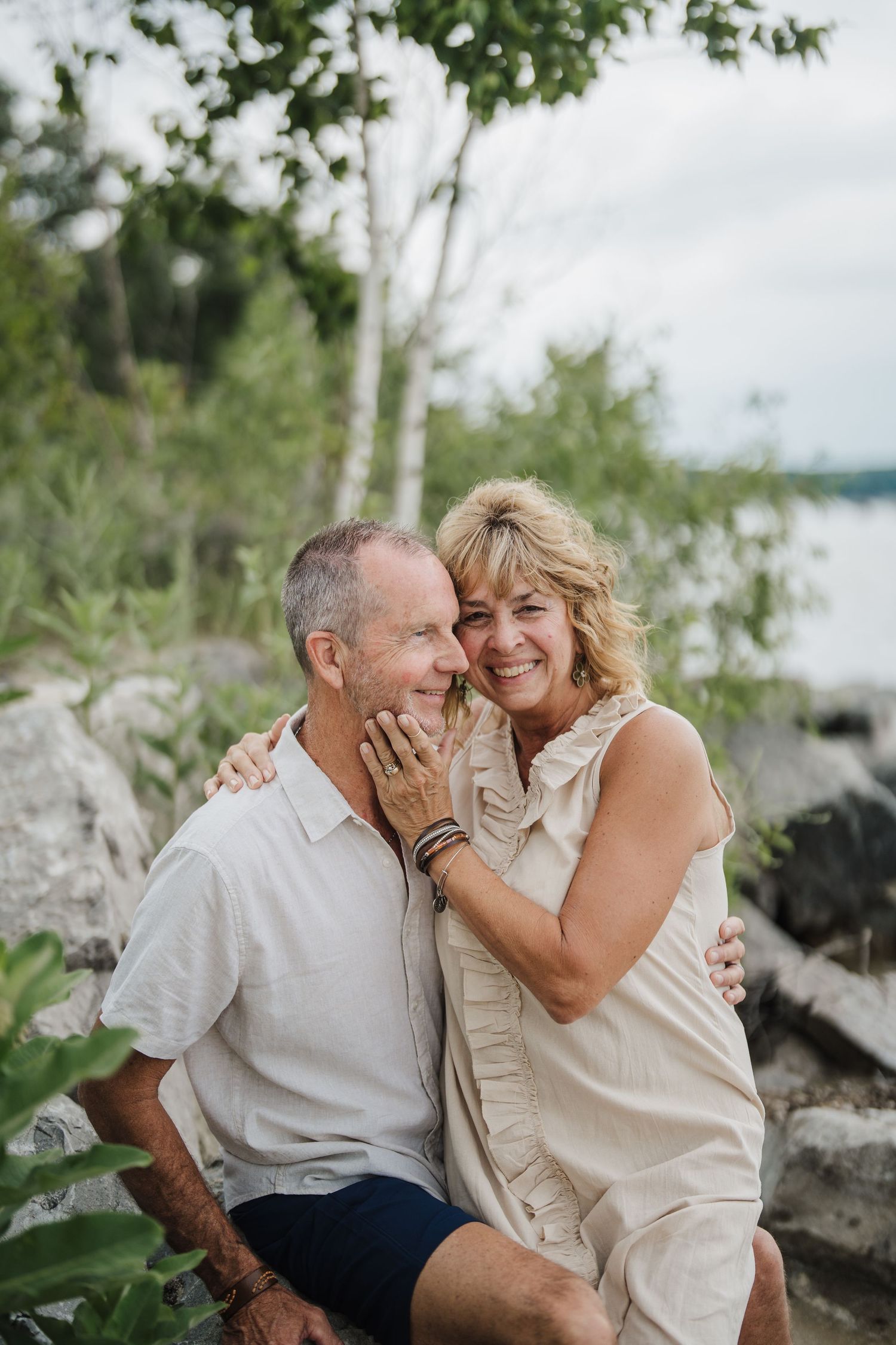 Couple sharing intimate laughing moments together on rocky shoreline with trees in background.