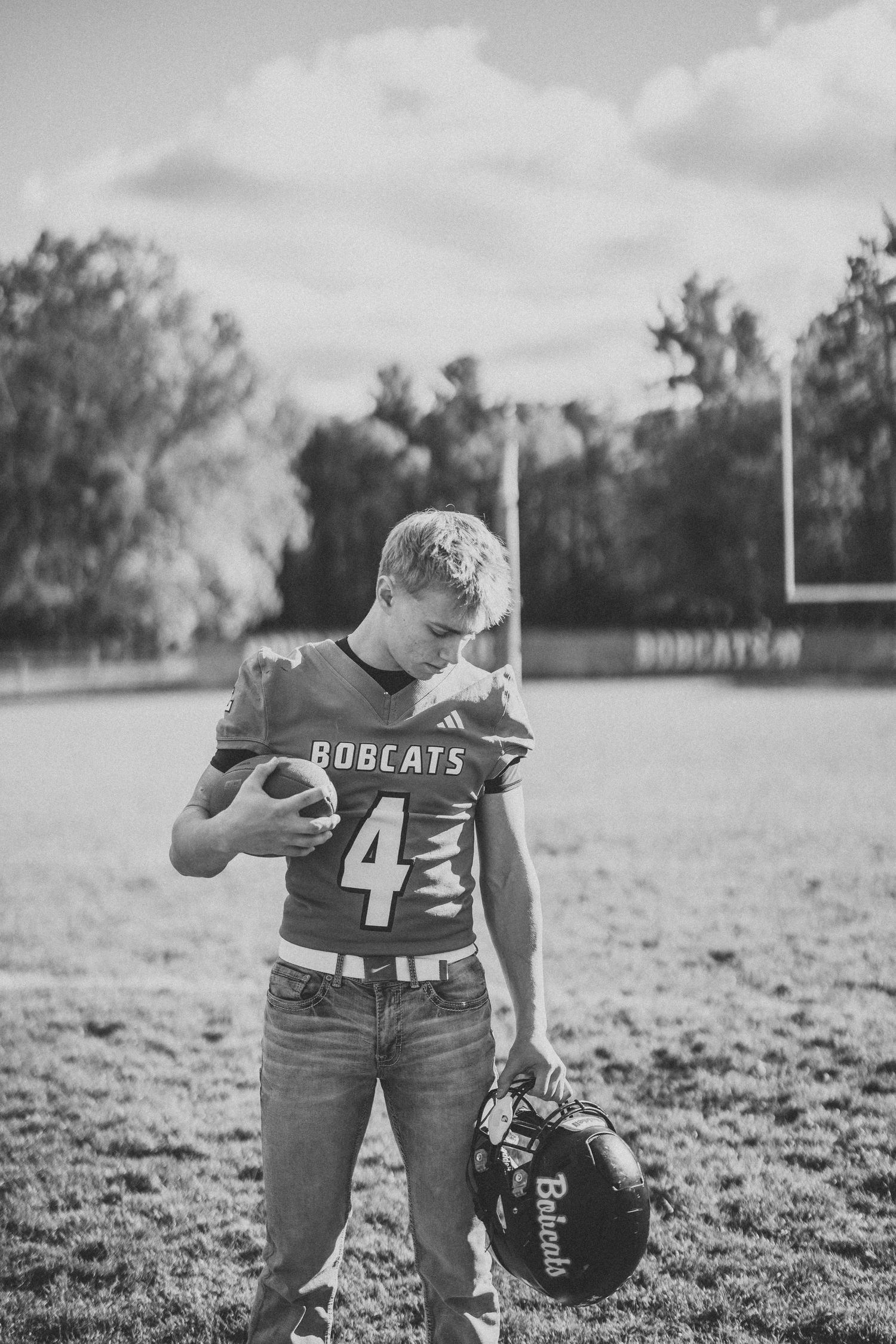 Person in football uniform poses with helmet in black and white on grassy field.